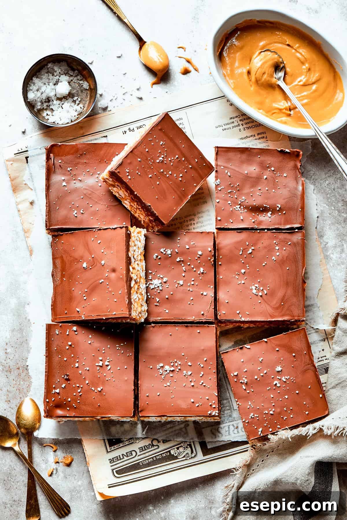 A delightful spread of golden peanut butter rice crispy treats, neatly cut into squares, invitingly displayed alongside bowls of creamy peanut butter and flaky sea salt. An array of freshly cut peanut butter rice crispy treats, perfectly arranged on a white surface, with a small bowl of creamy peanut butter and a pinch of sea salt nearby.