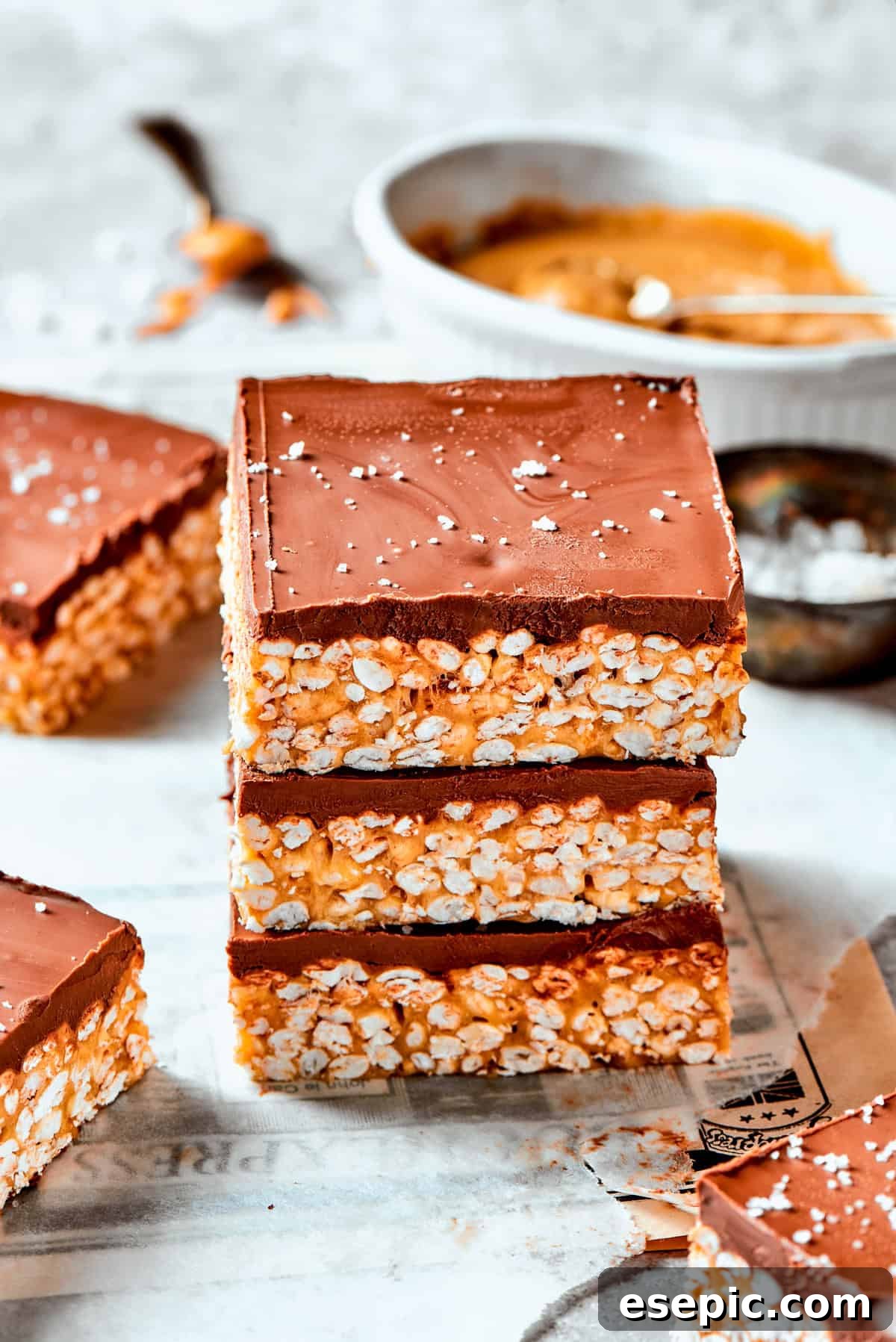 A tempting display of three golden peanut butter rice crispy bars, stacked gracefully, each boasting a luscious chocolate top and a tantalizingly chewy center. Three perfectly formed crispy rice bars, each adorned with a smooth layer of chocolate and a sprinkle of sea salt, are artfully stacked one on top of the other, showcasing their inviting texture.