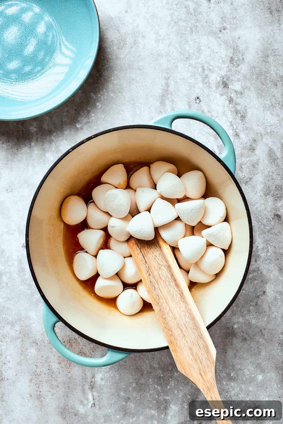 Mini marshmallows are added to the warm butter mixture, gradually melting into a smooth, sticky concoction. Fluffy white mini marshmallows are being stirred into the warm, melted butter mixture, beginning to soften and combine.