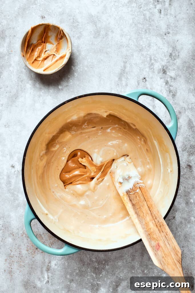 Peanut butter is carefully folded into the warm marshmallow mixture, ensuring every part of the base is rich with flavor. Creamy peanut butter is being incorporated into the melted marshmallow mixture, swirling together to create a rich, golden base.