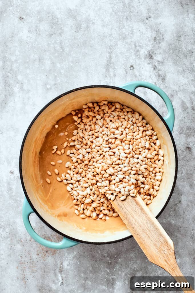 The crisp rice cereal is gently folded into the luscious peanut butter marshmallow blend, creating the beloved texture of the treats. Crisp rice cereal is being gently stirred into the thick, peanut butter marshmallow mixture, ensuring every flake is thoroughly coated.
