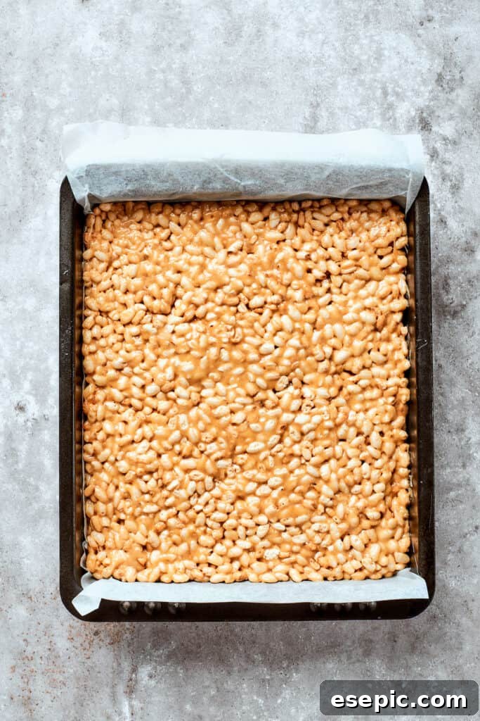 The rice crispy base is carefully pressed into a square dish, forming a smooth, uniform layer ready for its rich chocolate finish. The still-warm rice crispy treat mixture is being pressed firmly and evenly into a baking pan lined with parchment paper, forming a compact base.