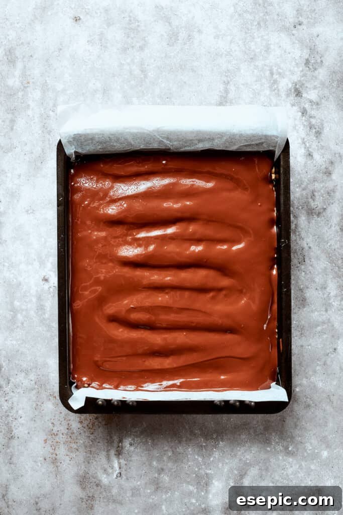 A luxurious blanket of melted chocolate is spread over the crispy rice base, promising a decadent finish once set. A gleaming, smooth layer of melted chocolate coats the pressed rice crispy treat base in a baking pan, ready to set.