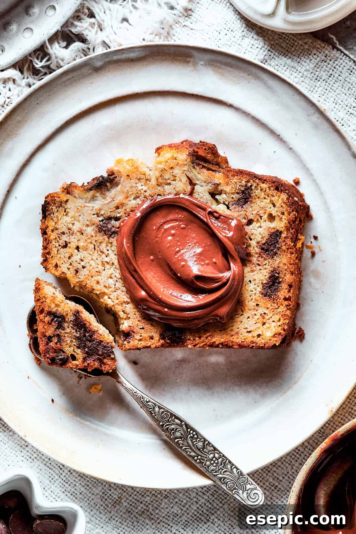 A hand gently spreading rich Nutella over a thick slice of warm, moist banana bread, promising a decadent experience.