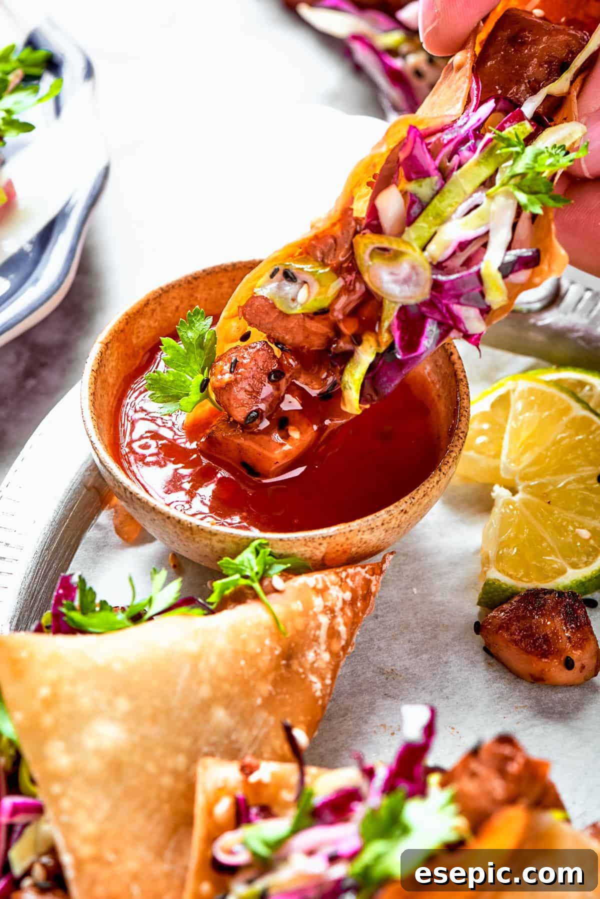 A hand dipping a crispy chicken wonton taco into a small bowl of vibrant red sweet chili sauce.