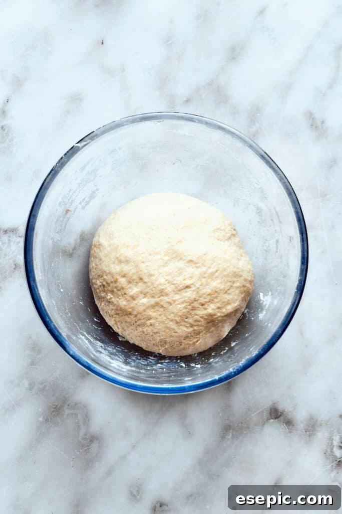 A perfectly smooth, elastic deep dish pizza dough ball rests in a glass bowl, covered and ready for its rise.