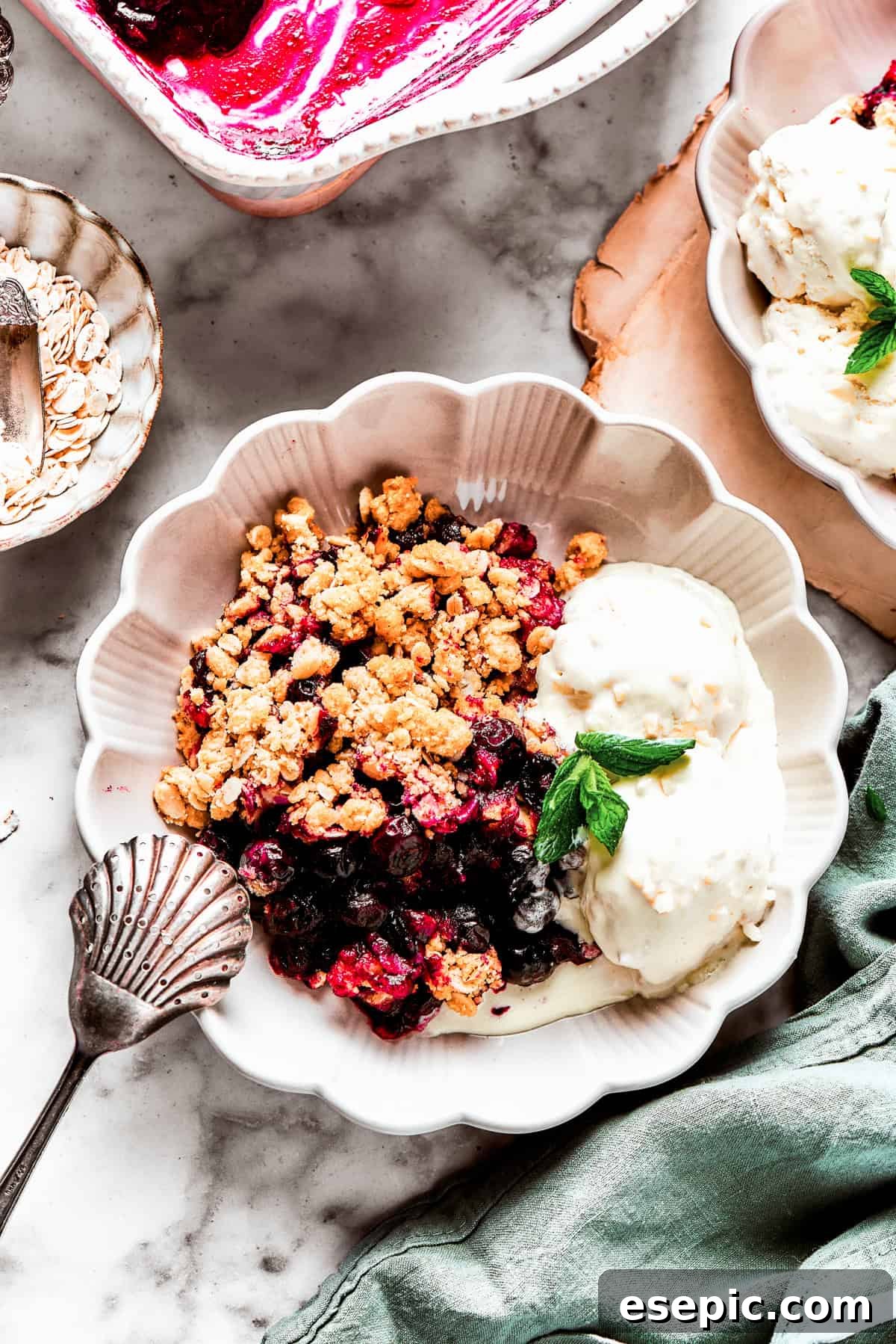 A perfect dessert: Blueberry crisp warm from the oven, topped with melting vanilla ice cream and a sprig of mint. A beautiful bowl of warm blueberry crisp, adorned with two scoops of vanilla ice cream that are starting to melt, and a vibrant sprig of fresh mint.