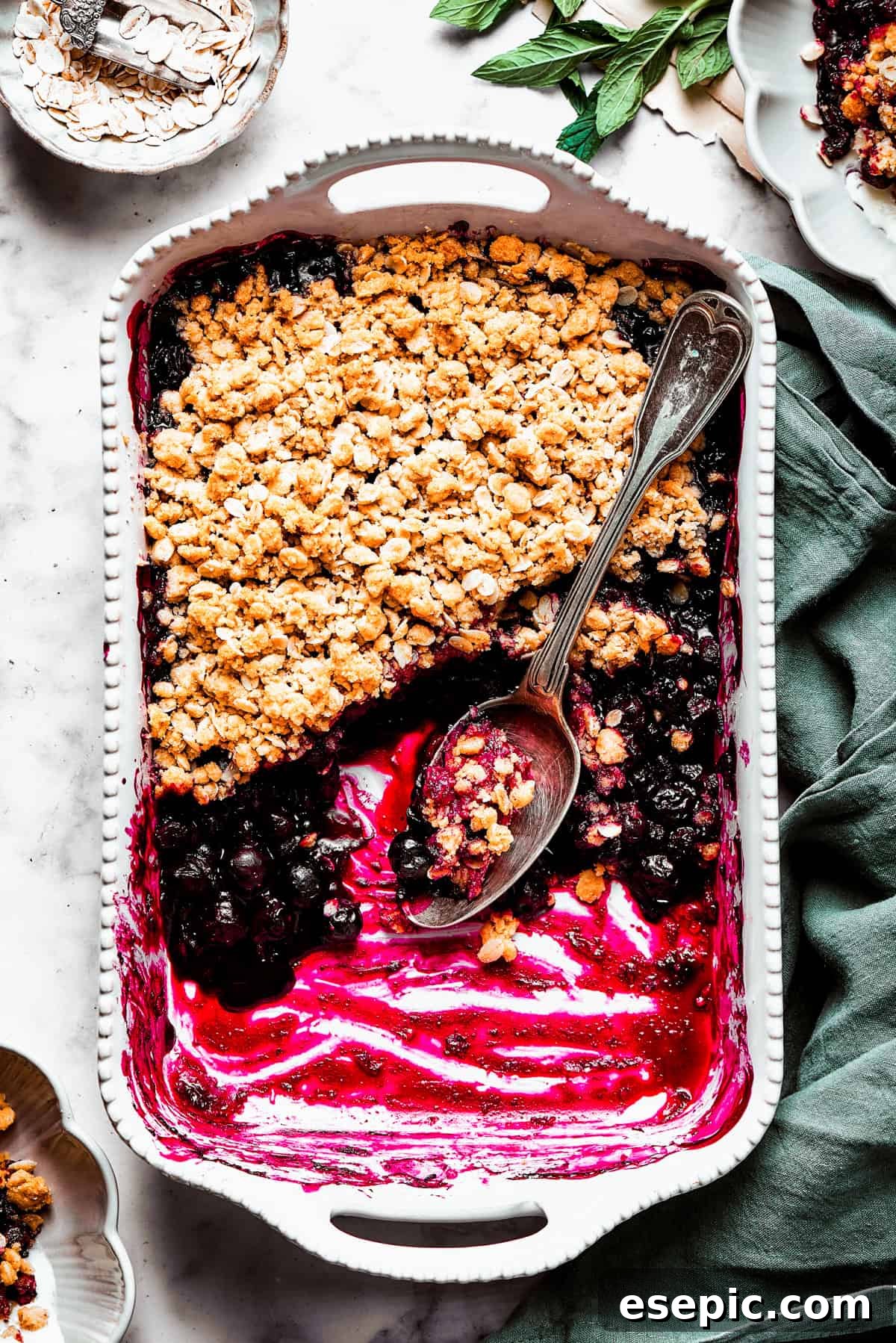 The ultimate comfort dessert: a warm blueberry crisp with a perfect portion missing, hinting at its deliciousness. An overhead view of a freshly baked blueberry crisp in a ceramic baking dish, with a portion scooped out, revealing the bubbly fruit filling beneath the golden crumble.