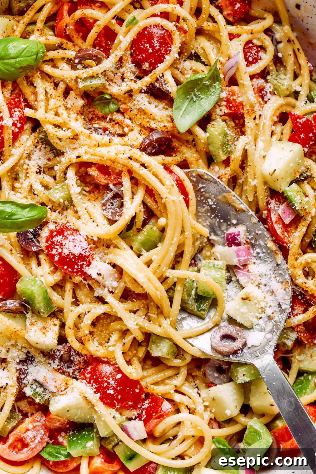 A close-up shot of spaghetti salad, highlighting the texture of the noodles, generous amounts of cheese, fresh vegetables like olives and bell peppers, and the rich dressing coating everything.