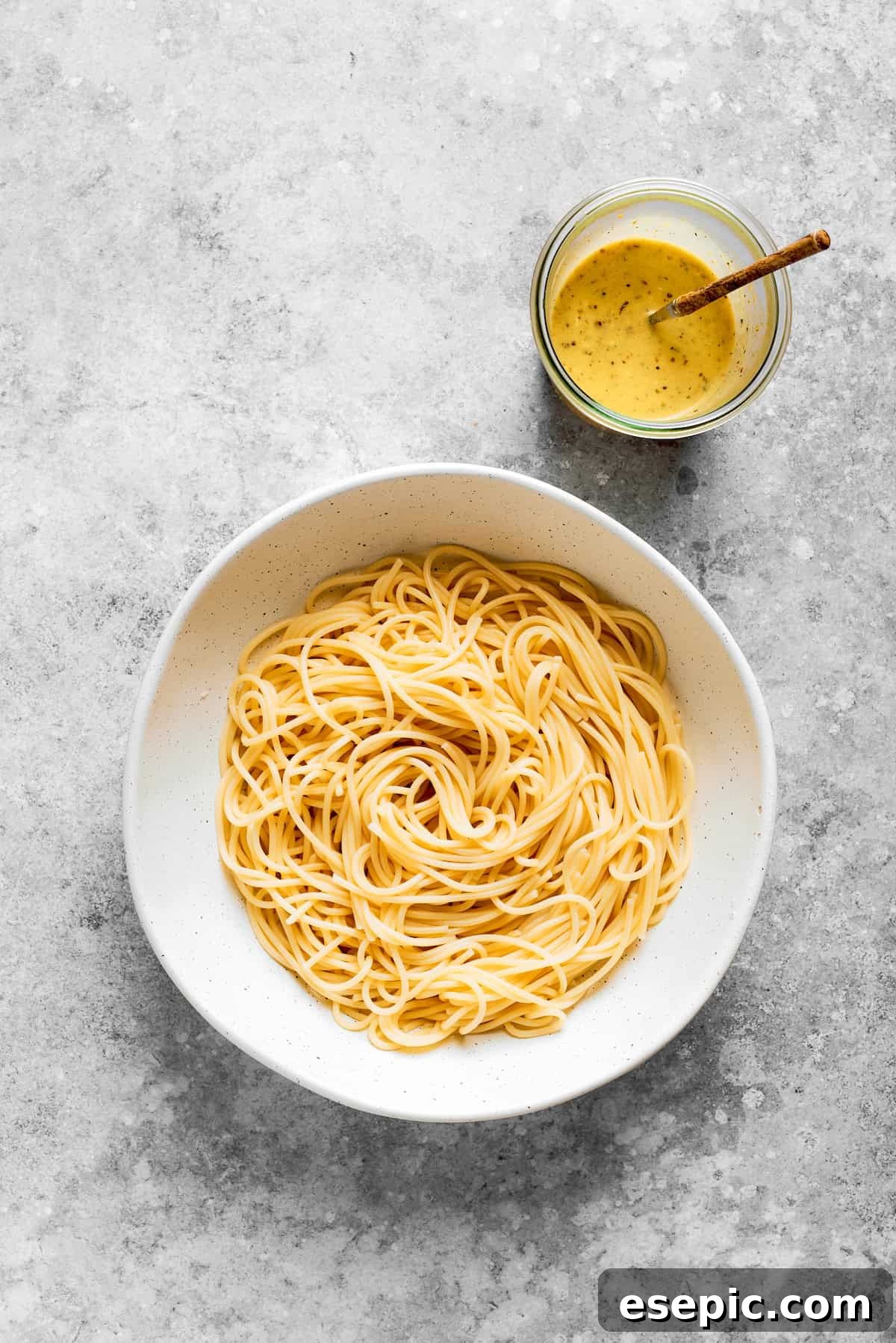 Cooked spaghetti noodles placed in a large white serving bowl, with a small bowl of dressing positioned beside it, ready to be combined.