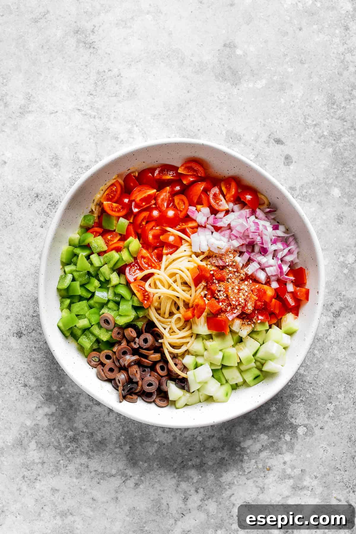 Freshly chopped vegetables, including bell peppers, tomatoes, and cucumbers, being added to a bowl of dressed spaghetti noodles, ready for the final toss.