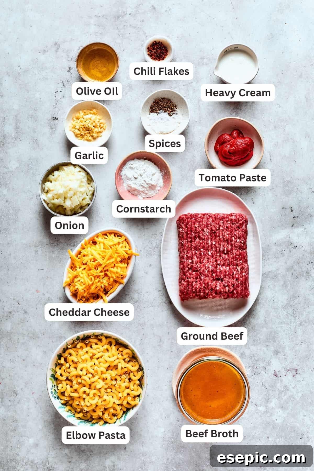 Hamburger helper ingredients laid out: ground meat, cheddar cheese, macaroni pasta, onion, garlic, cornstarch, tomato puree, olive oil, and heavy cream, all ready for cooking.