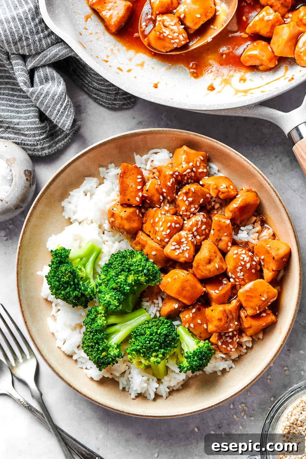 This Teriyaki Chicken Recipe folds tender bites of chicken into a thick, homemade teriyaki sauce for a savory and flavorful dish. A bowl of rice is topped with teriyaki chicken and broccoli with more teriyaki chicken in the background.
