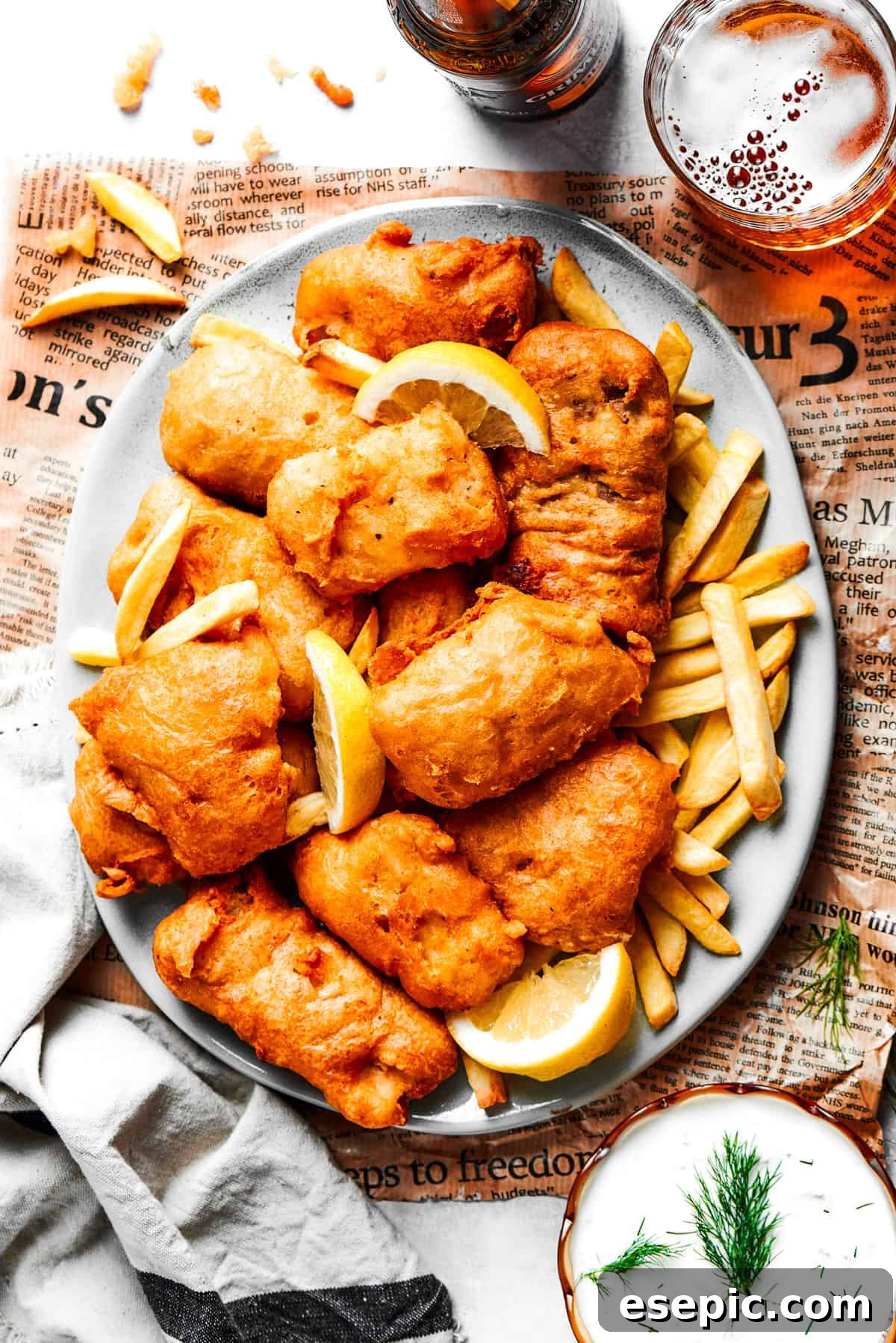 An inviting overhead shot of a generous platter piled high with golden-brown beer-battered fish and crispy French fries, with lemon wedges and tartar sauce on the side.