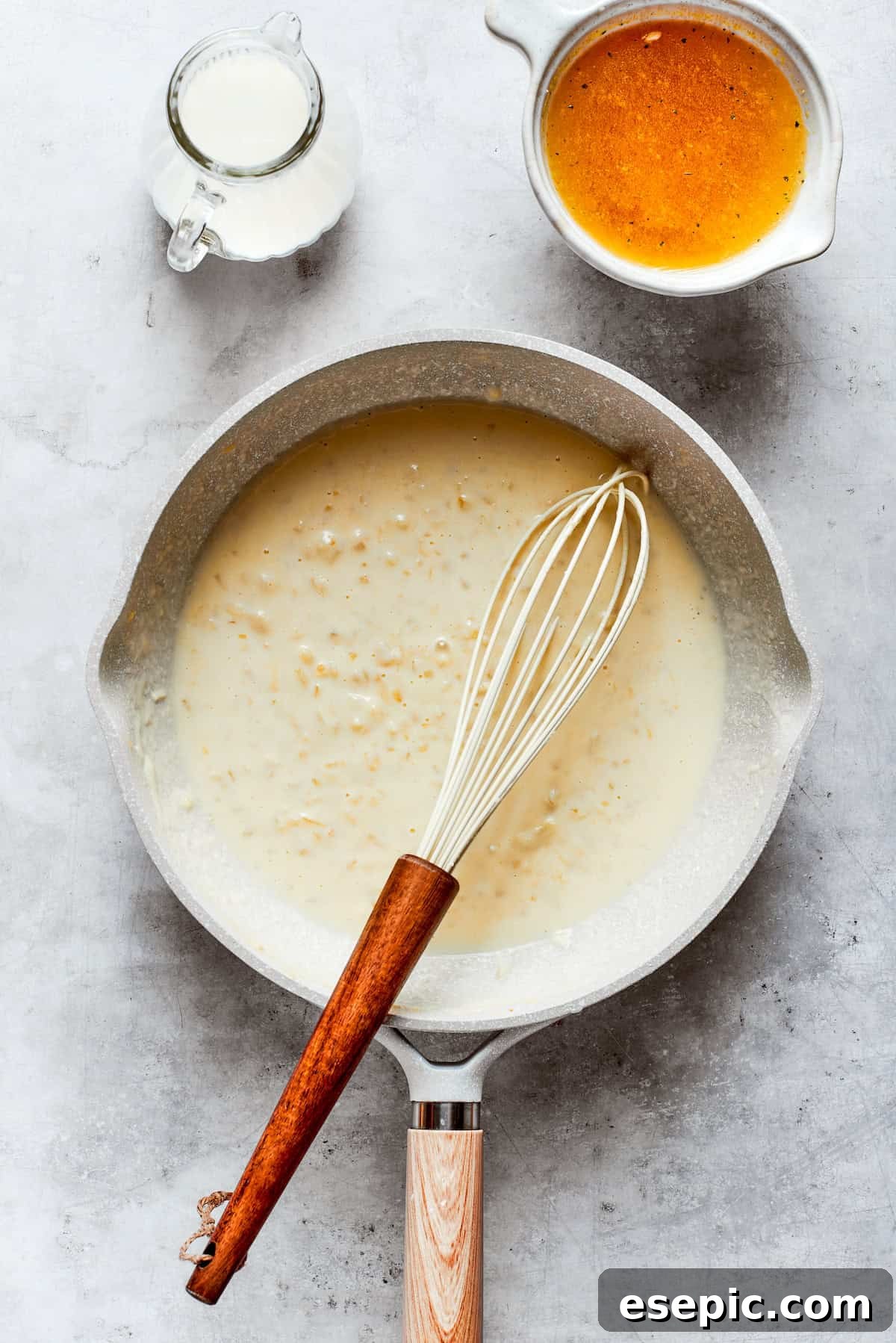 A whisk smoothly incorporates milk and broth into the roux in a pot, beginning to form the creamy cheese sauce.