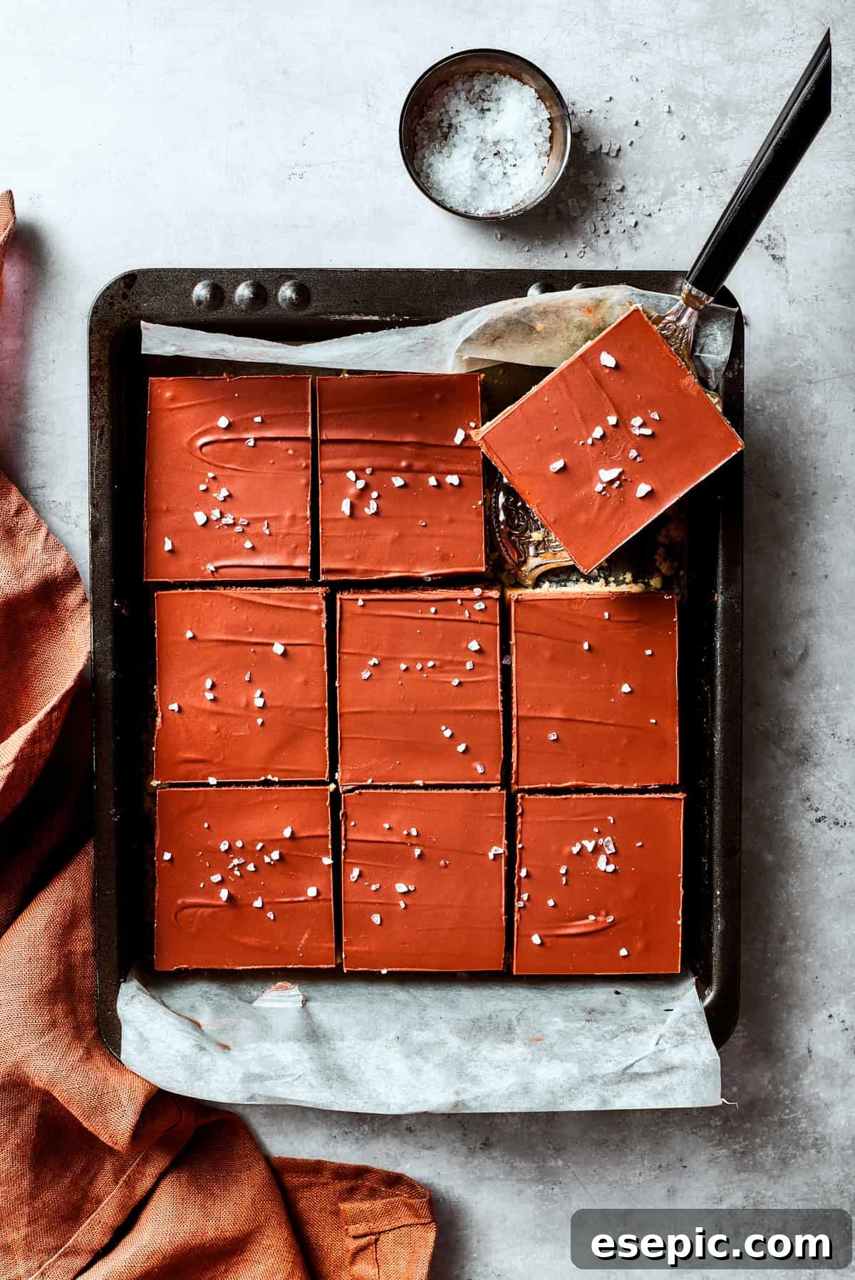 Millionaire Bars are decadent layers of homemade buttery shortbread, creamy caramel, and thick chocolate ganache. A serving utensil gently lifting a perfectly cut square of Millionaire Bar from the pan, showcasing its beautiful layers.