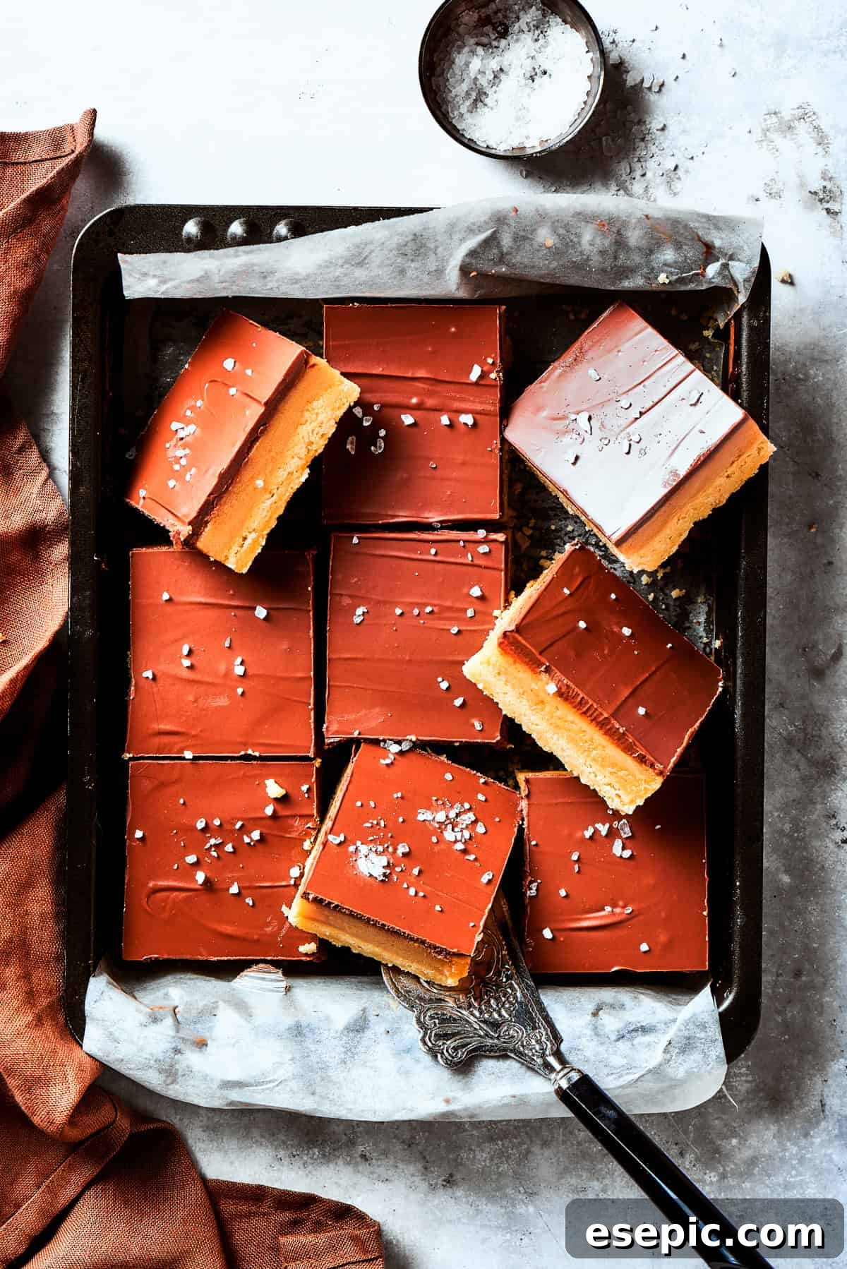 Millionaire Bars are decadent layers of homemade buttery shortbread, creamy caramel, and thick chocolate ganache. A square baking pan filled with freshly made Millionaire Bars, showcasing their elegant layers before cutting.