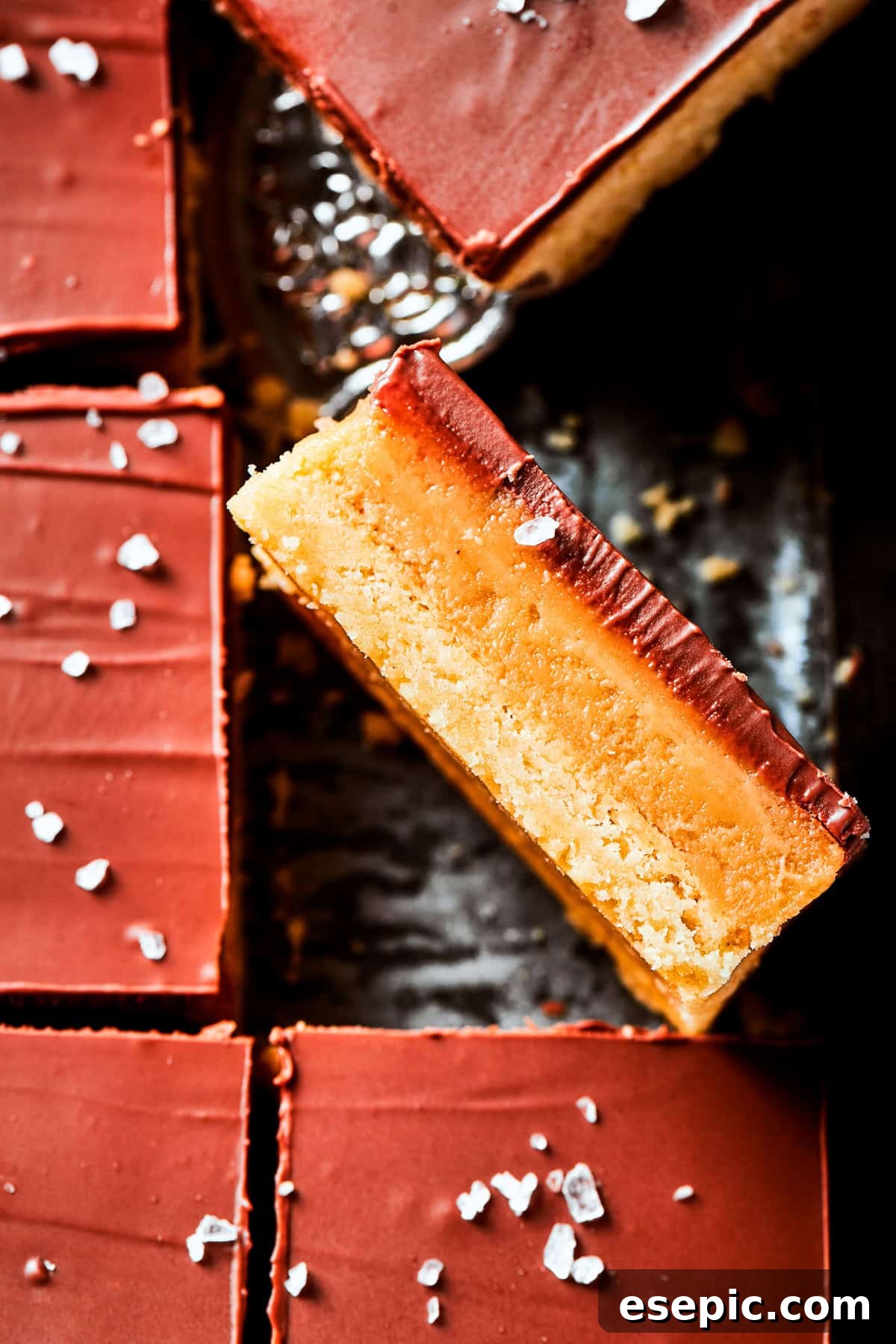 Millionaire Bars are decadent layers of homemade buttery shortbread, creamy caramel, and thick chocolate ganache. A single Millionaire Bar, cut and turned on its side, showcasing the distinct layers of shortbread, caramel, and chocolate, resting on a pan of uncut bars.