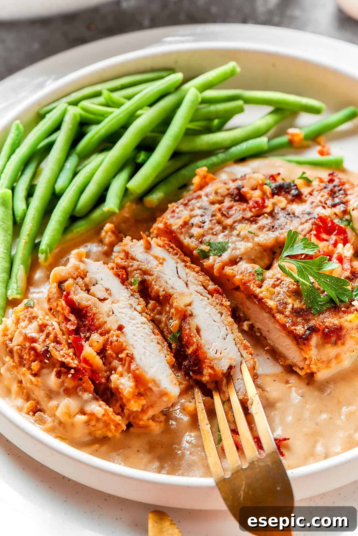 Smothered Chicken breasts feature pan-fried chicken in a crispy breadcrumb coating topped with a rich and creamy homemade gravy. A beautifully plated piece of smothered chicken, sliced to reveal its tender interior, served alongside vibrant green beans.