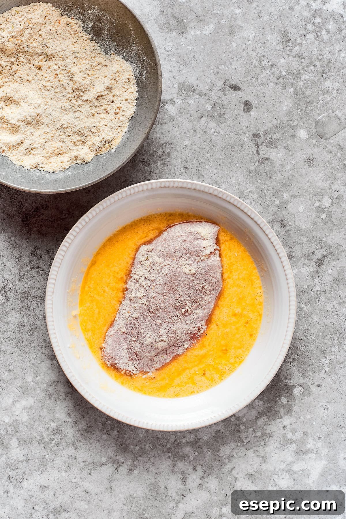 Smothered Chicken breasts feature pan-fried chicken in a crispy breadcrumb coating topped with a rich and creamy homemade gravy. A chicken breast is carefully dipped into a bowl of egg wash, ensuring full coverage.