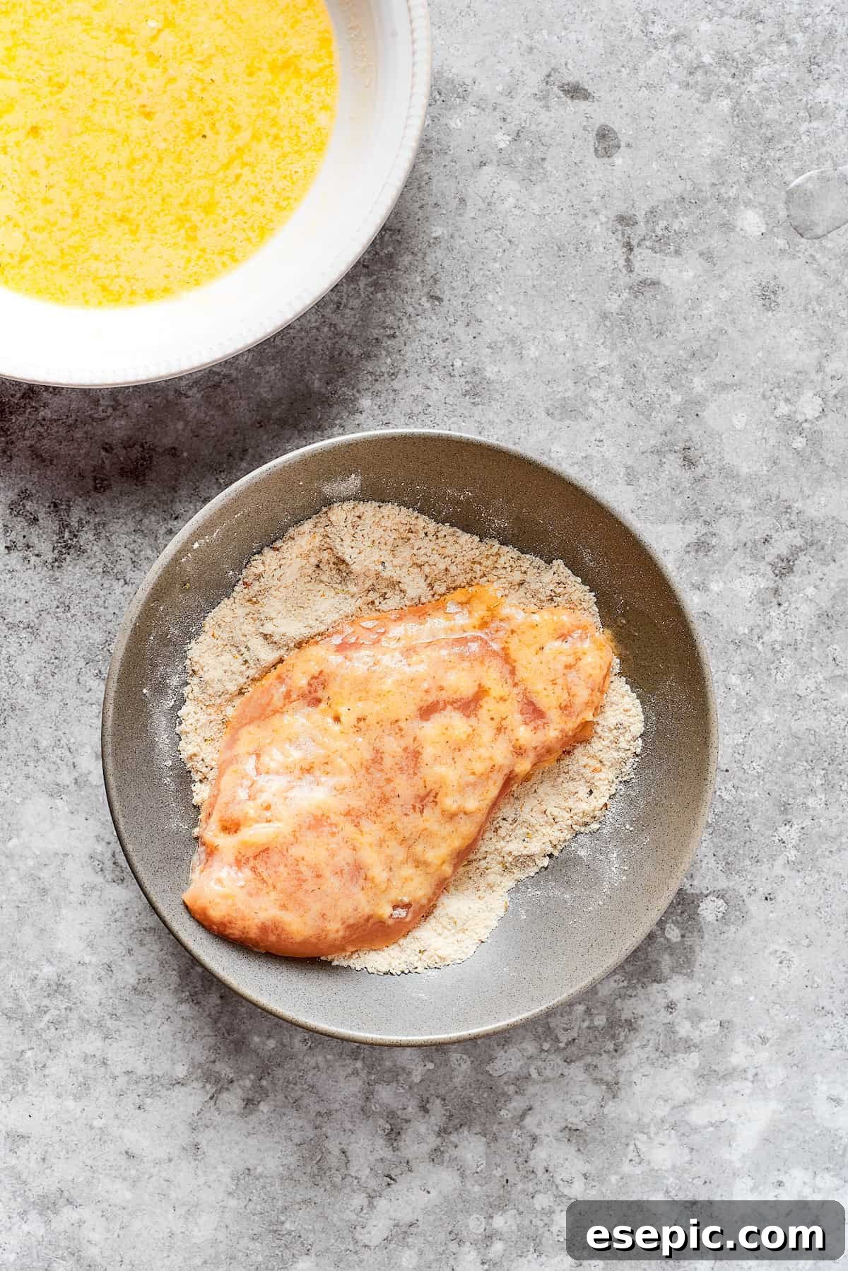 Smothered Chicken breasts feature pan-fried chicken in a crispy breadcrumb coating topped with a rich and creamy homemade gravy. A chicken breast is thoroughly dredged in a bowl of breadcrumbs, ensuring an even and complete coating.