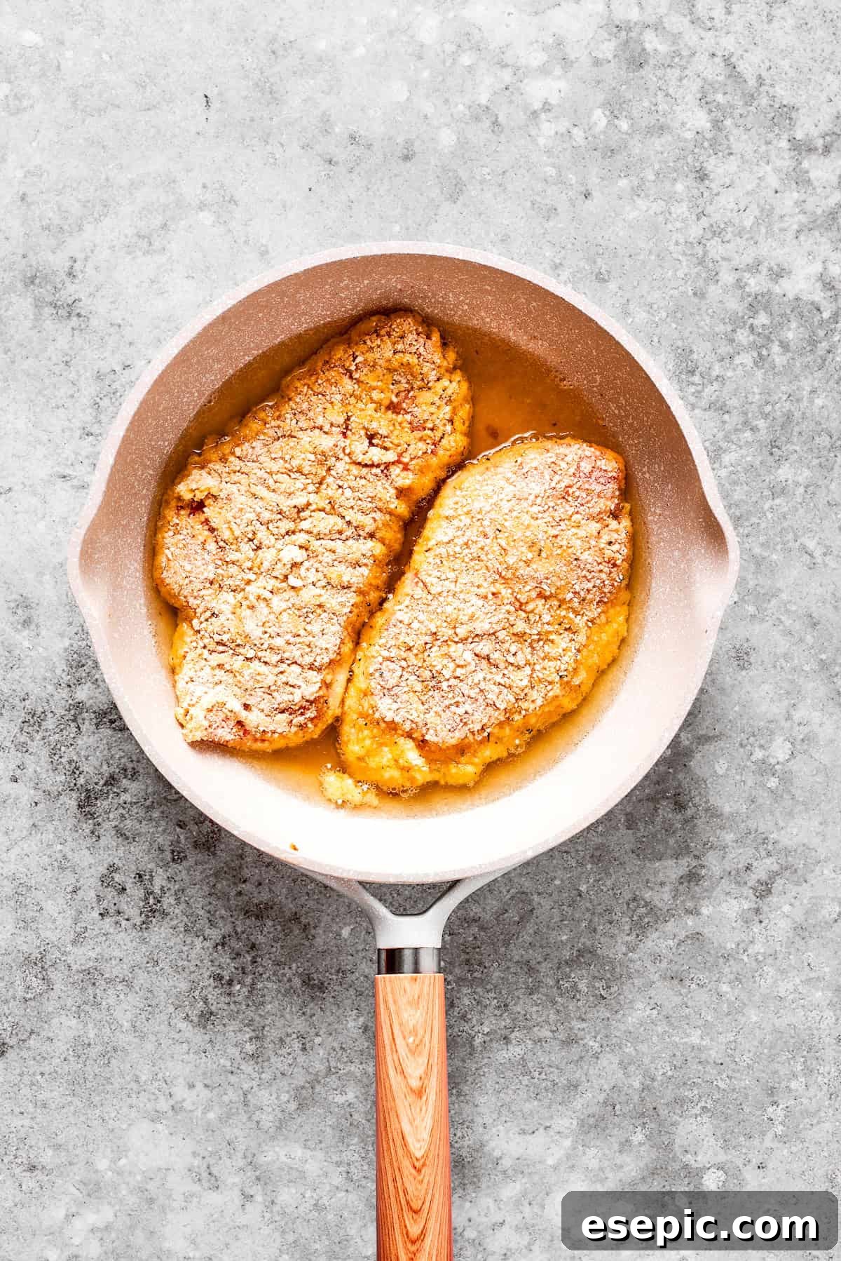 Smothered Chicken breasts feature pan-fried chicken in a crispy breadcrumb coating topped with a rich and creamy homemade gravy. Two breaded chicken breasts are pan-frying in a skillet, turning golden brown.