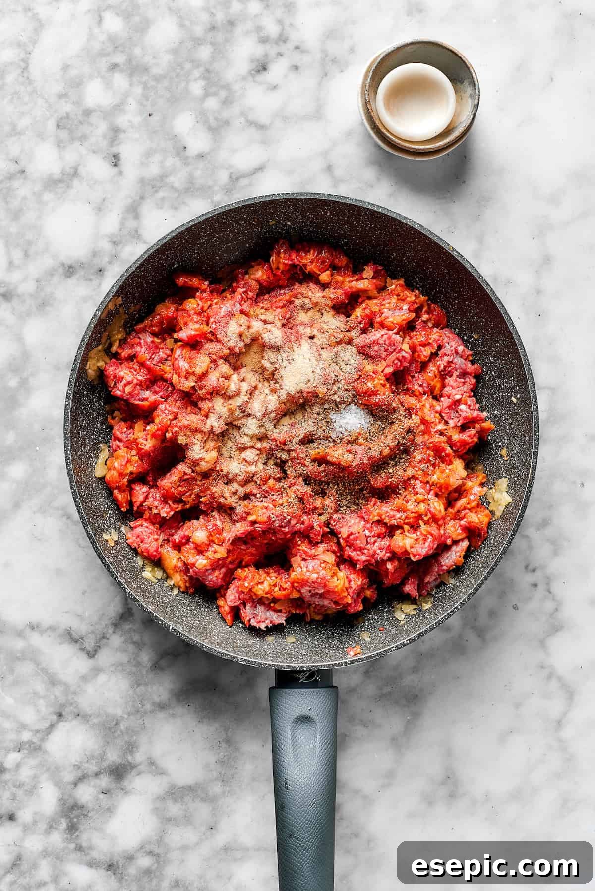 Seasonings being stirred into browned ground meat in a skillet.