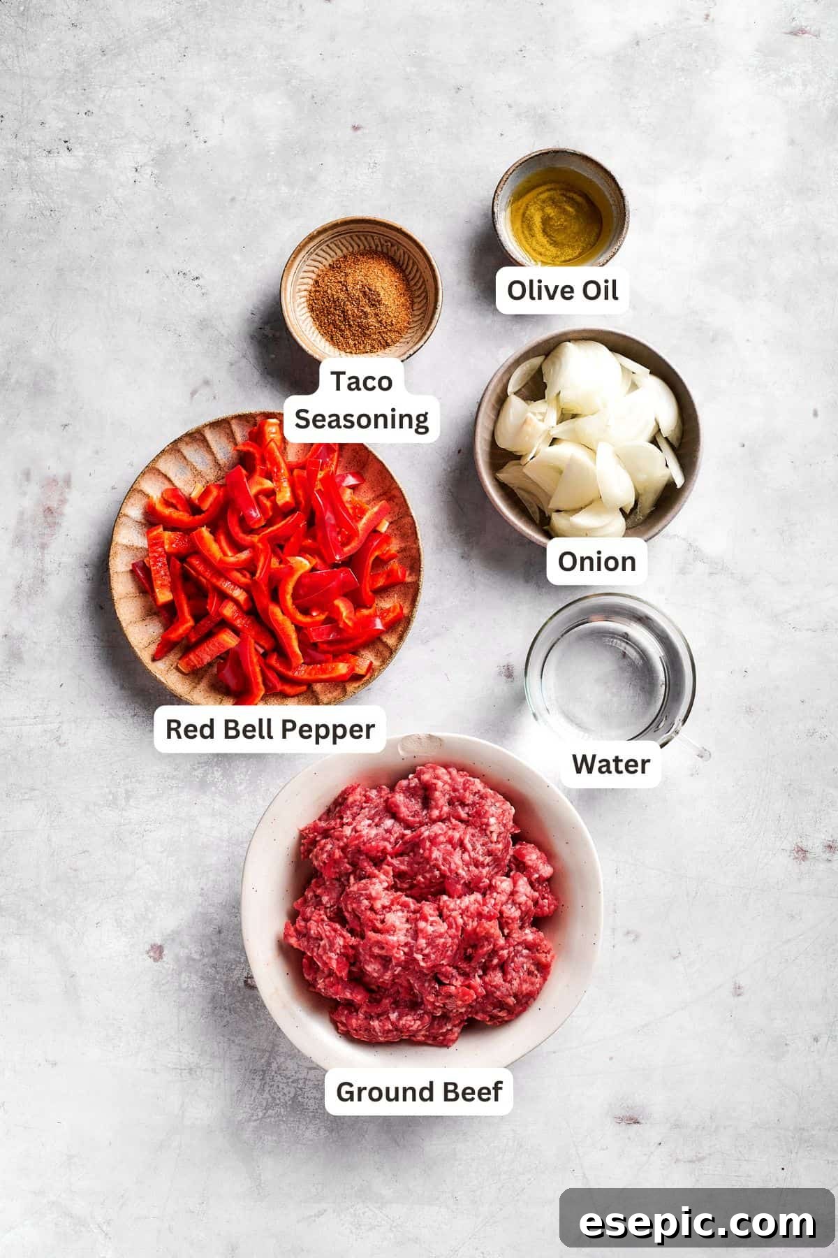 Assortment of fresh ingredients for John Wayne Casserole, including ground beef, sliced tomatoes, onions, bell peppers, cheese, sour cream, Bisquick mix, and taco seasoning, all neatly arranged in small bowls.