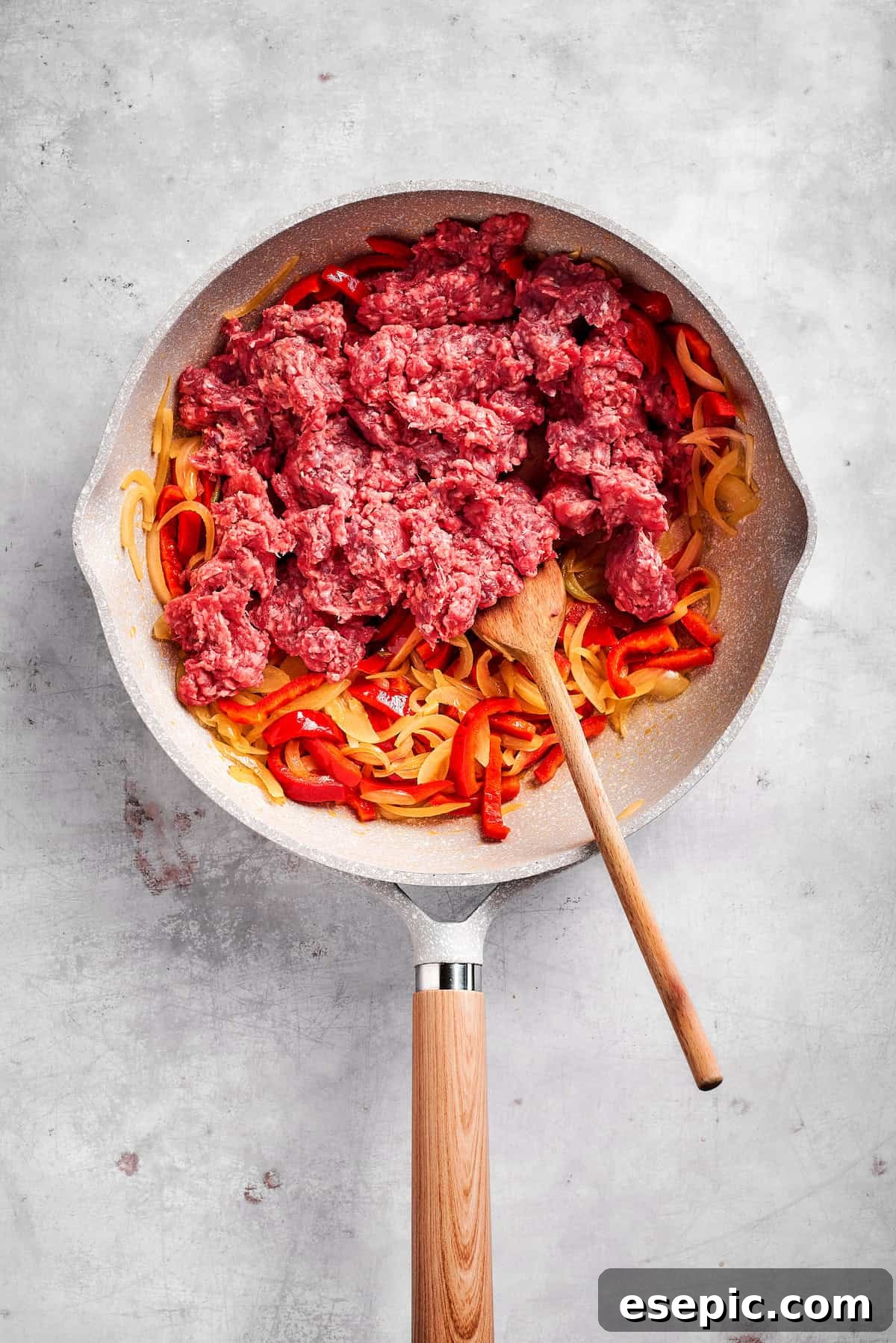 Ground beef being added to a skillet containing sautéed onions and bell peppers, ready to be browned for the casserole filling.