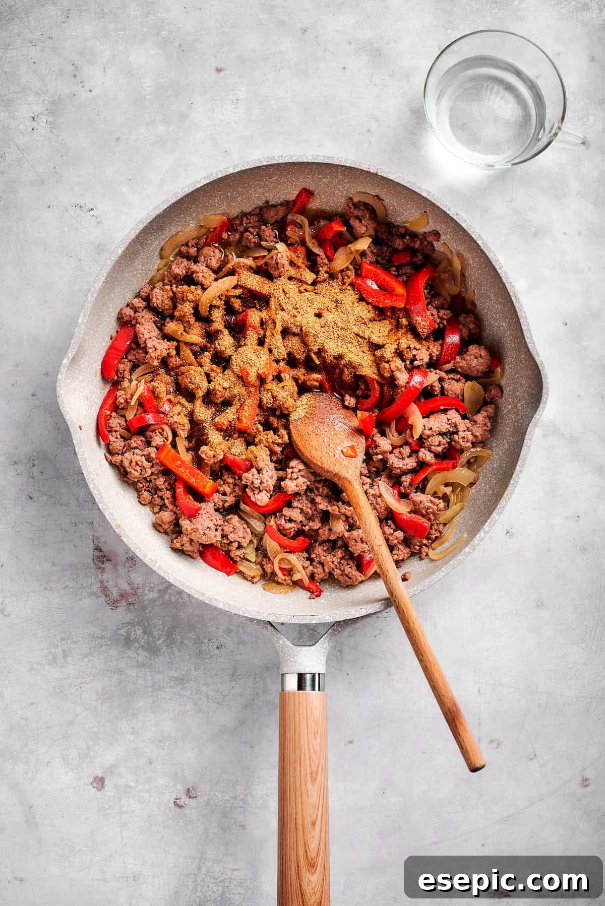 Taco seasoning being sprinkled over cooked ground beef and sautéed vegetables in a skillet, ready to be mixed for the casserole filling.