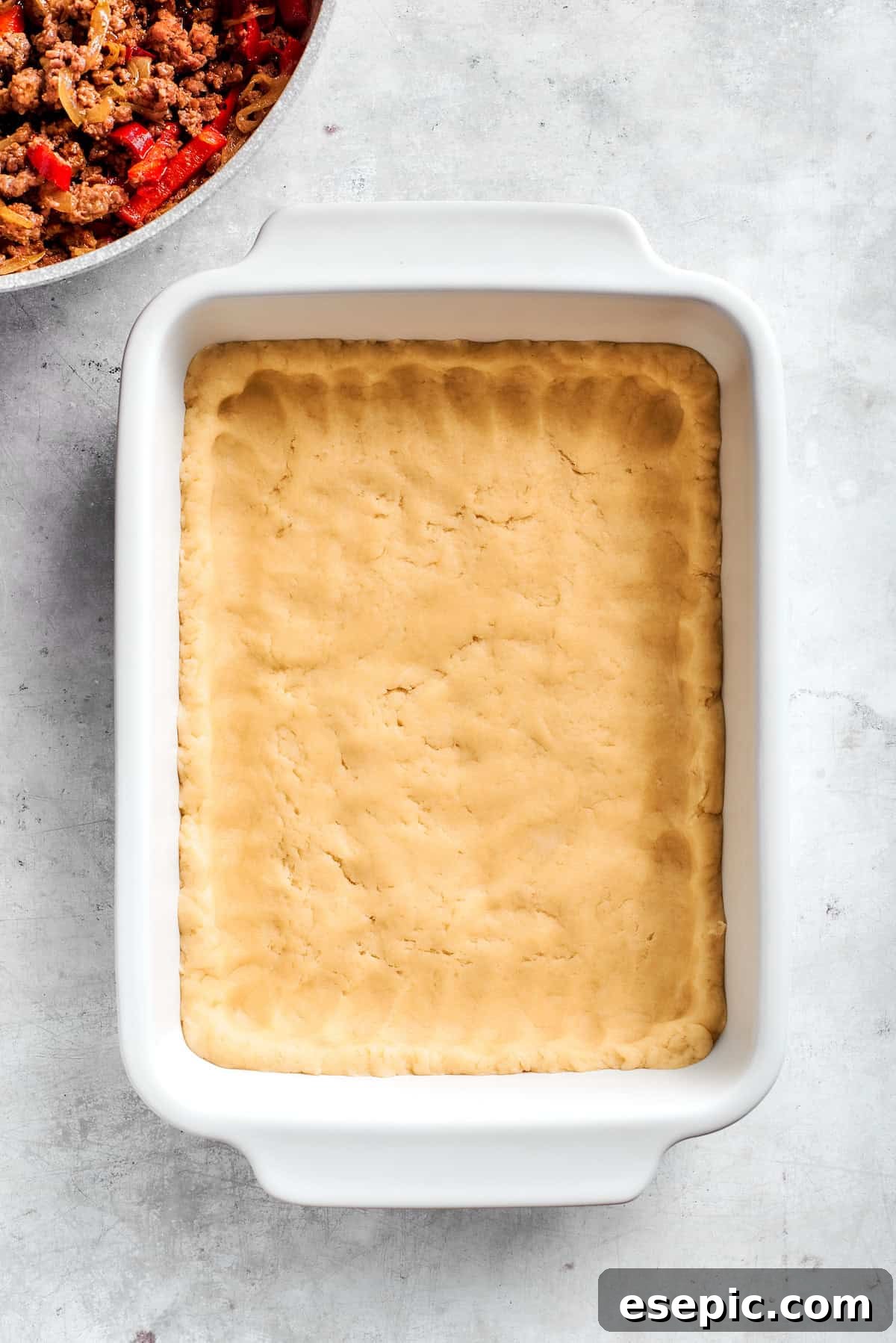 Biscuit dough being evenly pressed into the bottom of a rectangular baking dish, forming the base of the casserole.