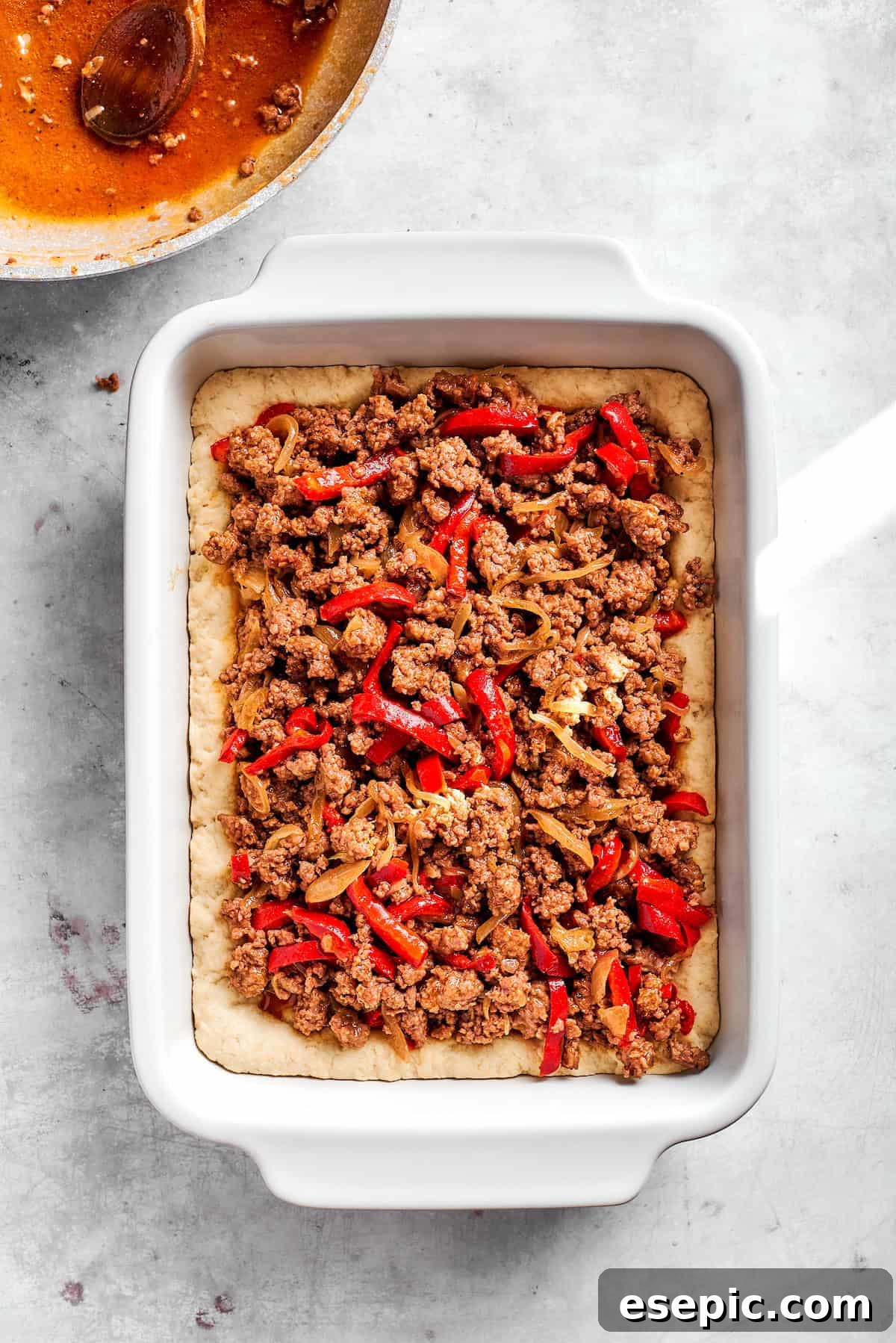 Cooked ground beef and vegetable filling being spread over the biscuit base in a baking dish, forming the next layer of the casserole.