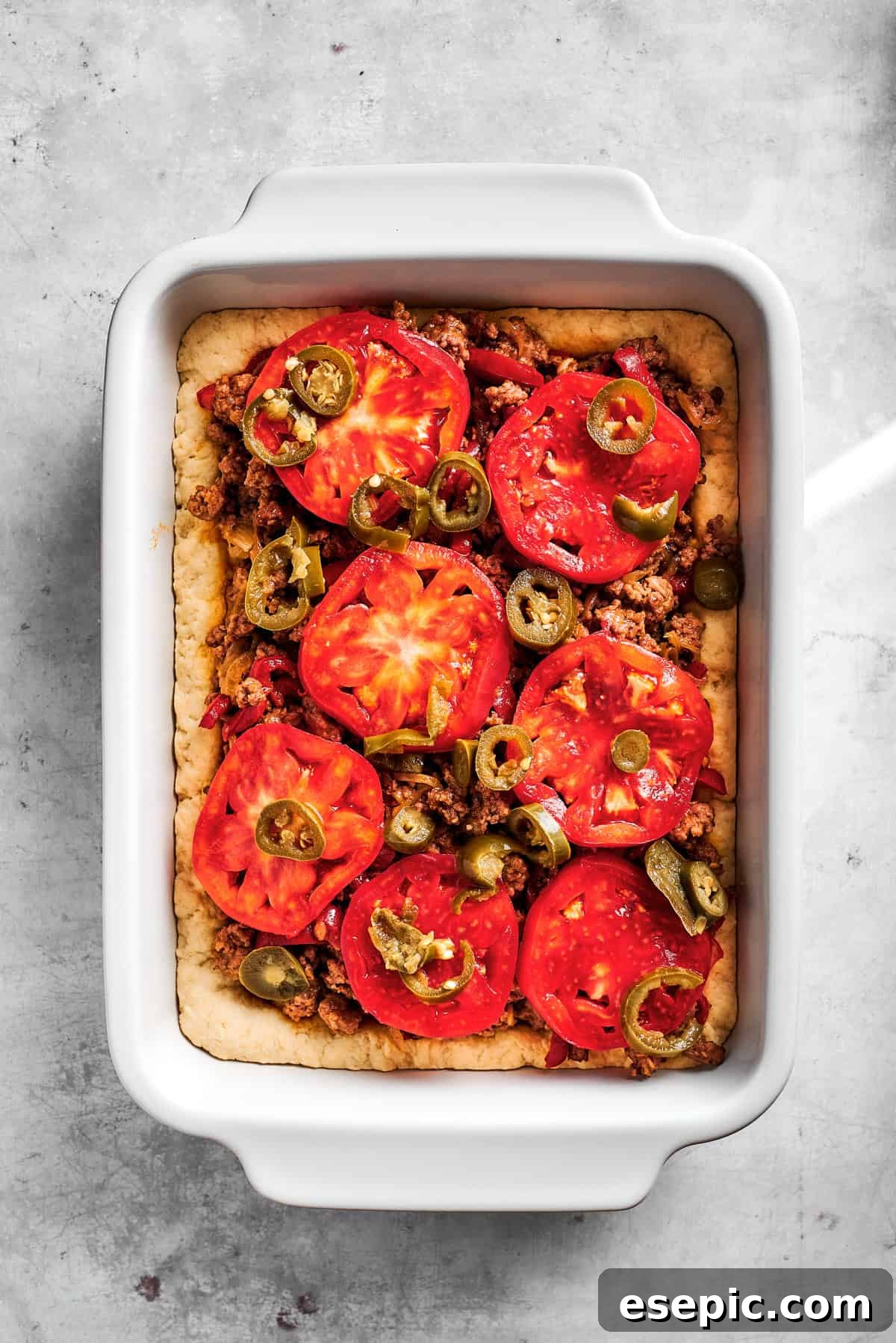 Thin slices of fresh Roma tomatoes arranged neatly over the ground beef layer in the baking dish, adding freshness and color to the casserole.