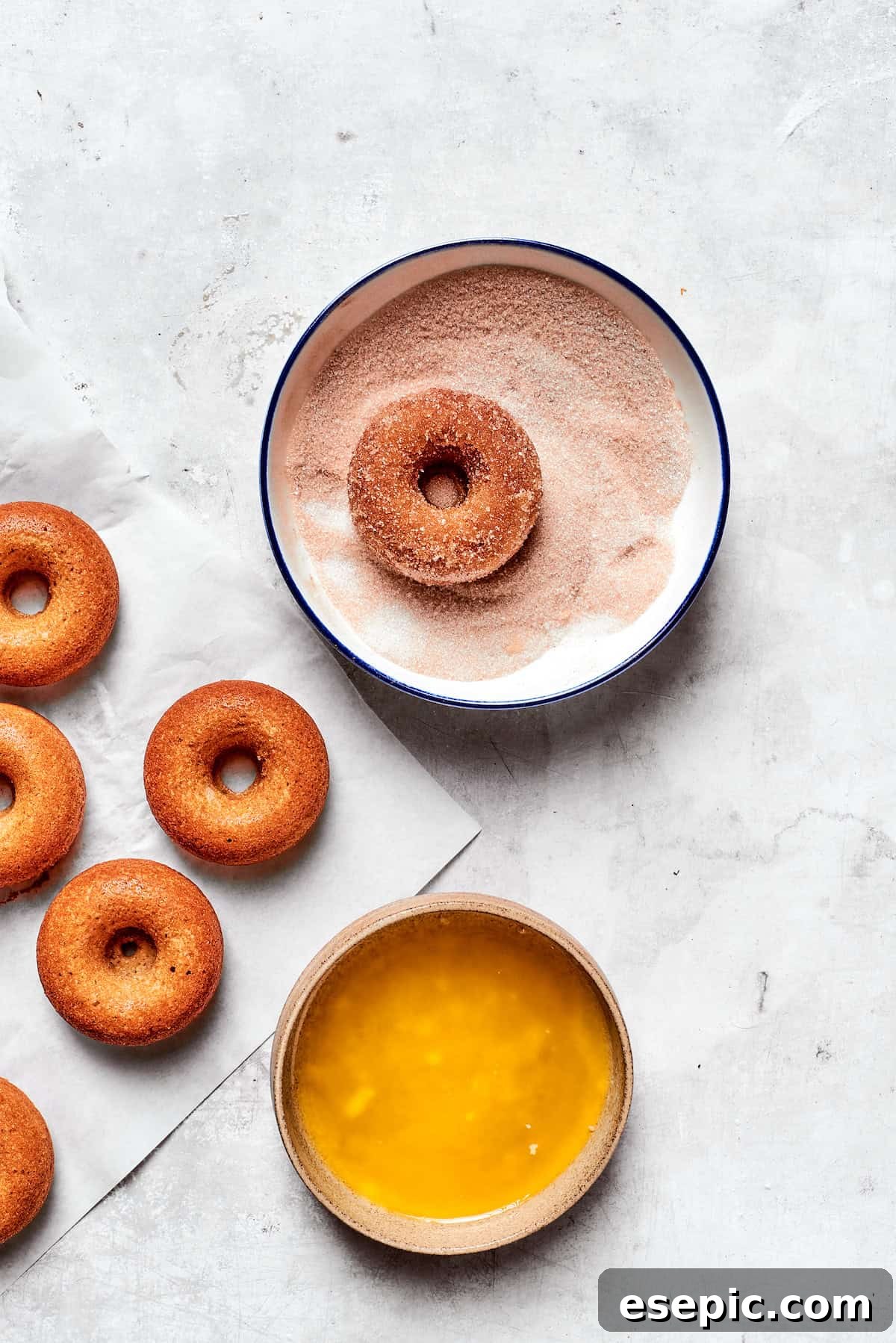 Baked not fried apple cider donuts are perfectly tender, dusted with spiced sugar, and fragrant with sweet apple cider. An apple cider donut being dipped into a bowl of cinnamon sugar after being brushed with melted butter, ensuring full coating.