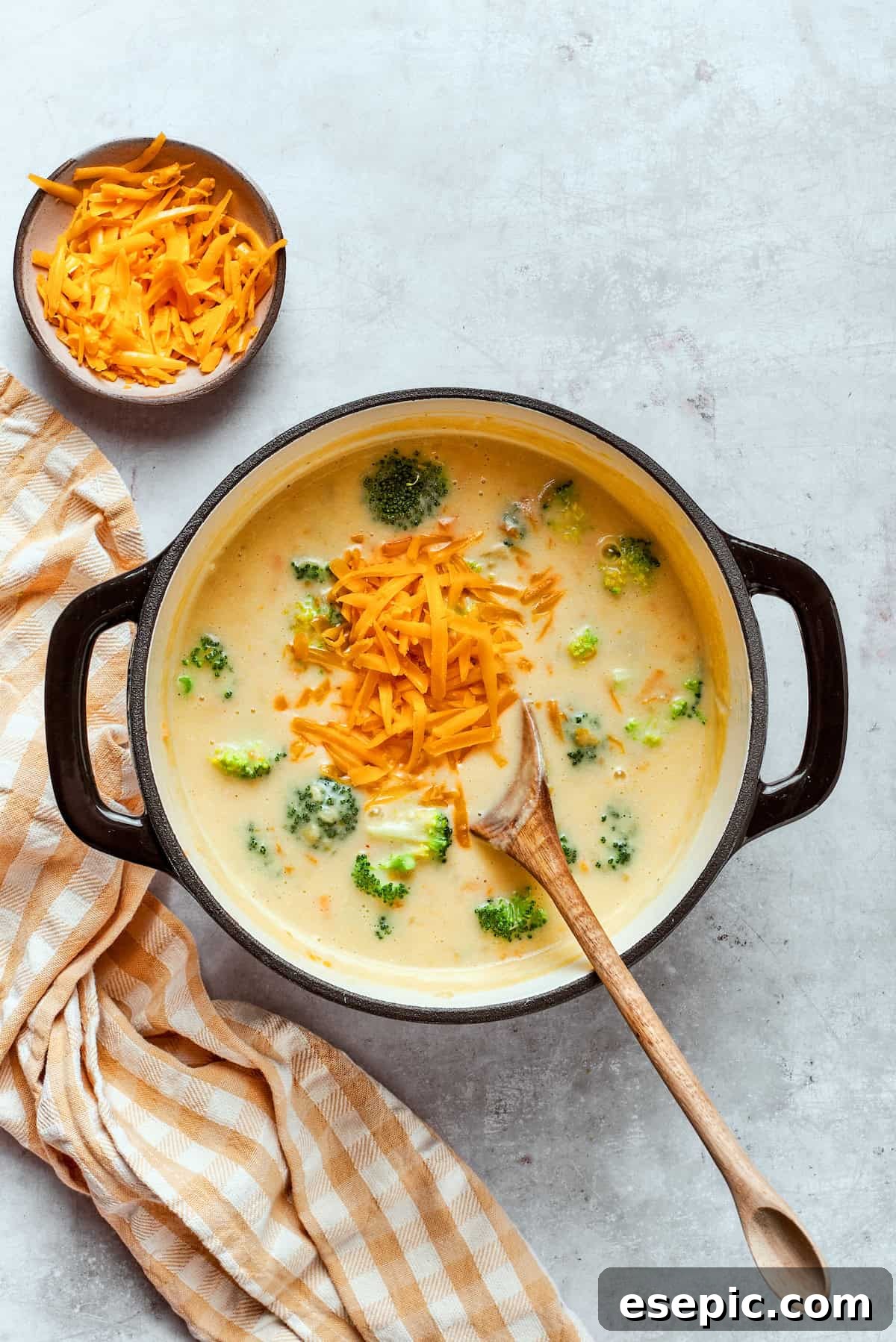 This easy broccoli cheddar soup recipe is packed with cozy flavor, featuring broccoli florets, diced potatoes, and freshly grated cheddar cheese. Adding shredded cheddar to a pot with soup.