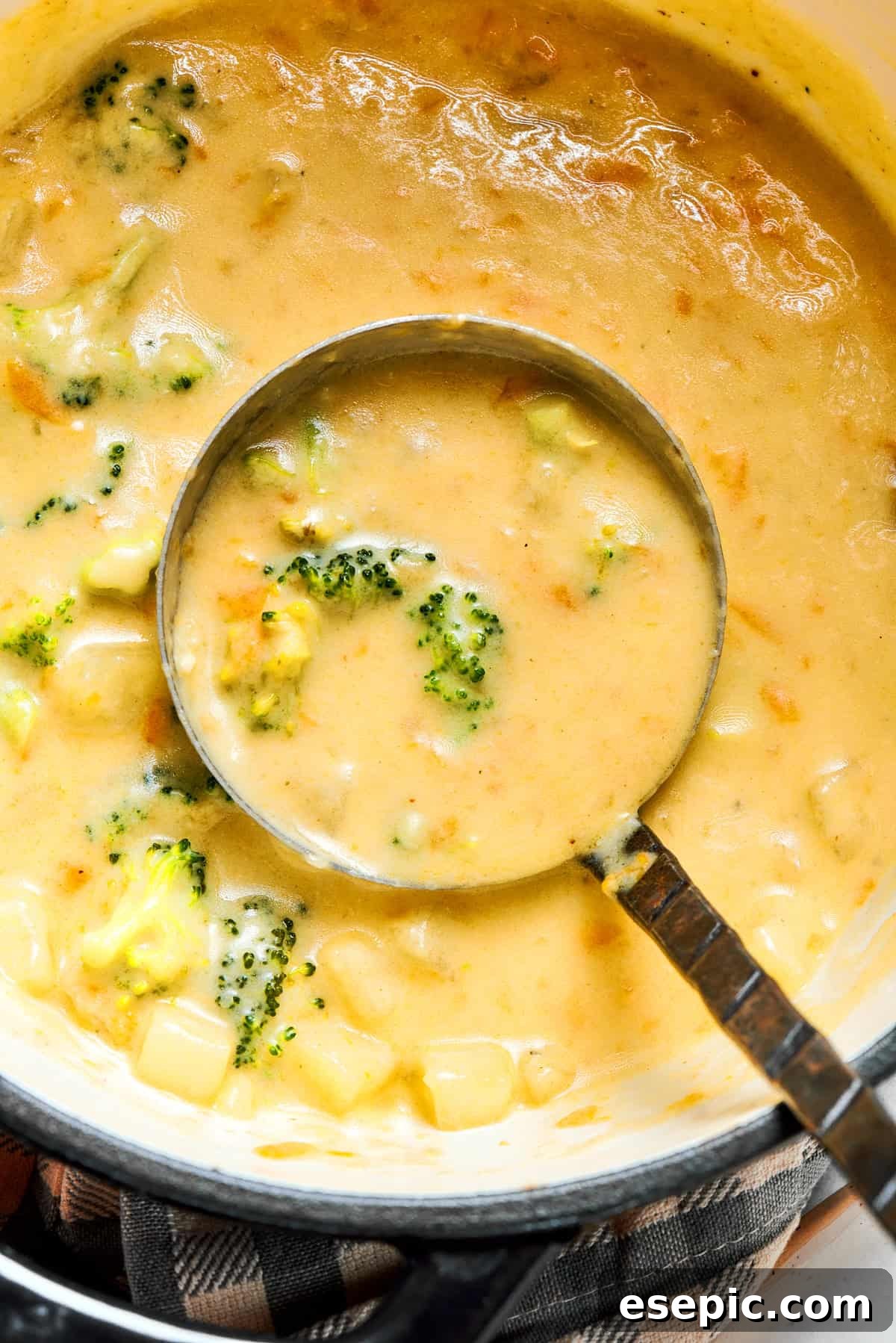This easy broccoli cheddar soup recipe is packed with cozy flavor, featuring broccoli florets, diced potatoes, and freshly grated cheddar cheese. A ladle gently spooning out a generous portion of creamy broccoli cheese soup from a large cooking pot.