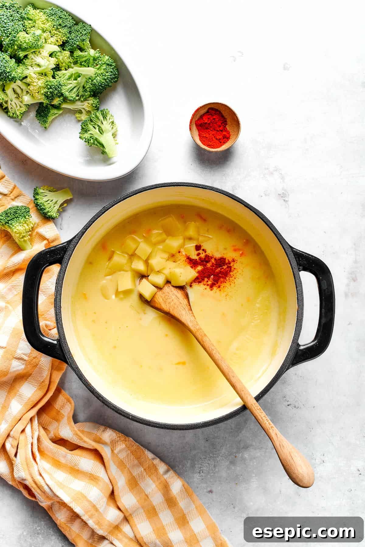 This easy broccoli cheddar soup recipe is packed with cozy flavor, featuring broccoli florets, diced potatoes, and freshly grated cheddar cheese. Adding diced potato and paprika to soup in a pot.