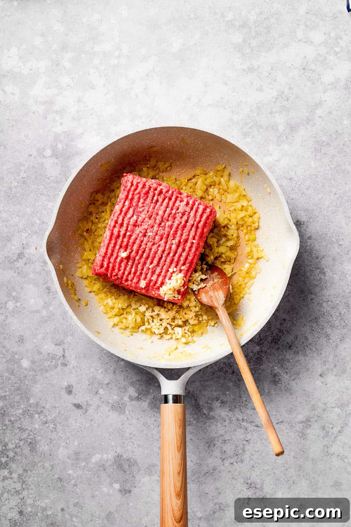 A large block of raw ground beef is carefully added to a hot frying pan with sautéed onions, ready to be browned.