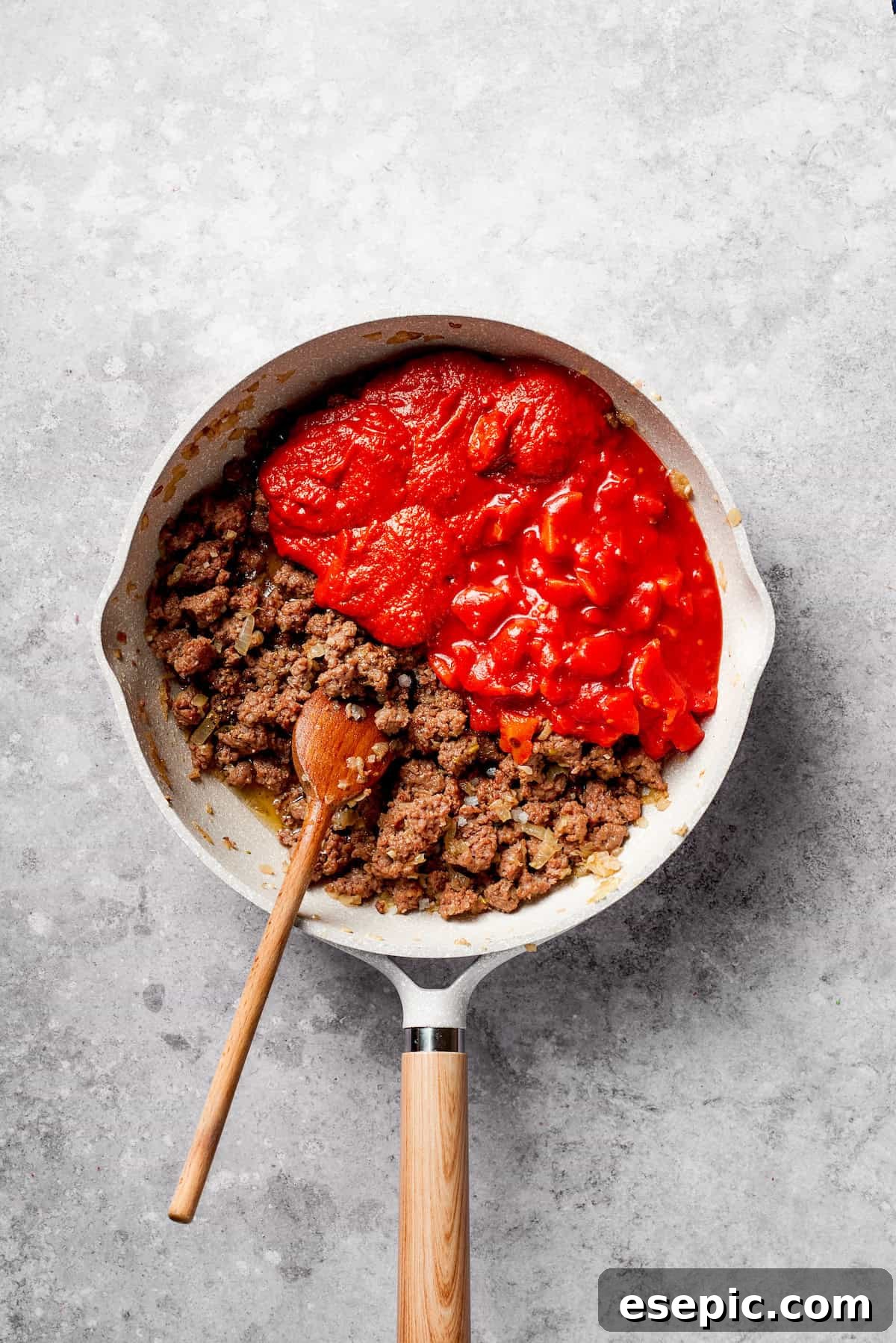 Rich canned tomatoes are stirred into the browned ground beef and onion mixture in a large frying pan, forming the base of the savory sauce.