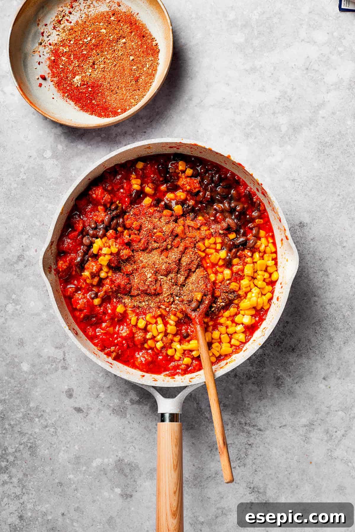 A vibrant mix of black beans and yellow corn is added to the simmering ground beef and tomato sauce in a pan, ready to be stirred for even distribution.