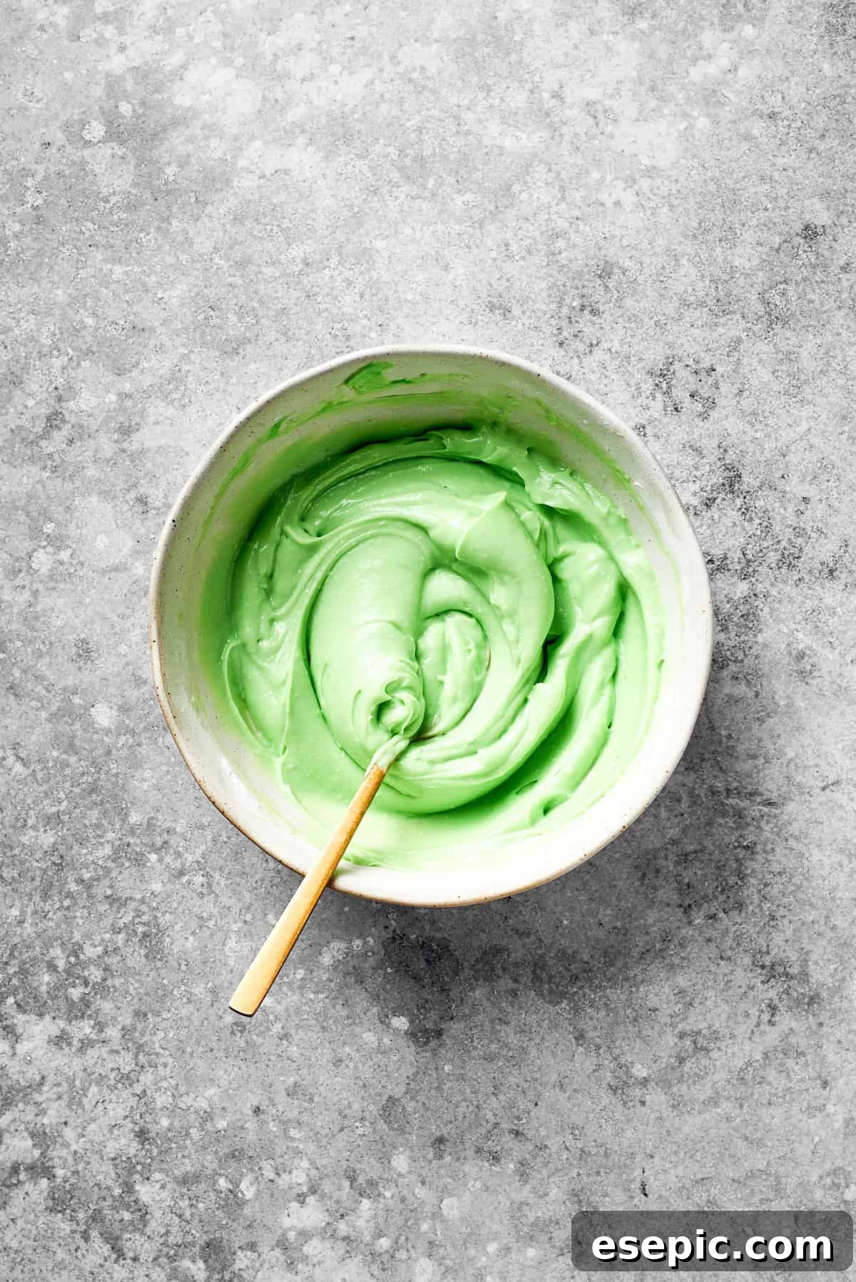 A spoon stirs green food coloring into icing, achieving a uniform vibrant green.