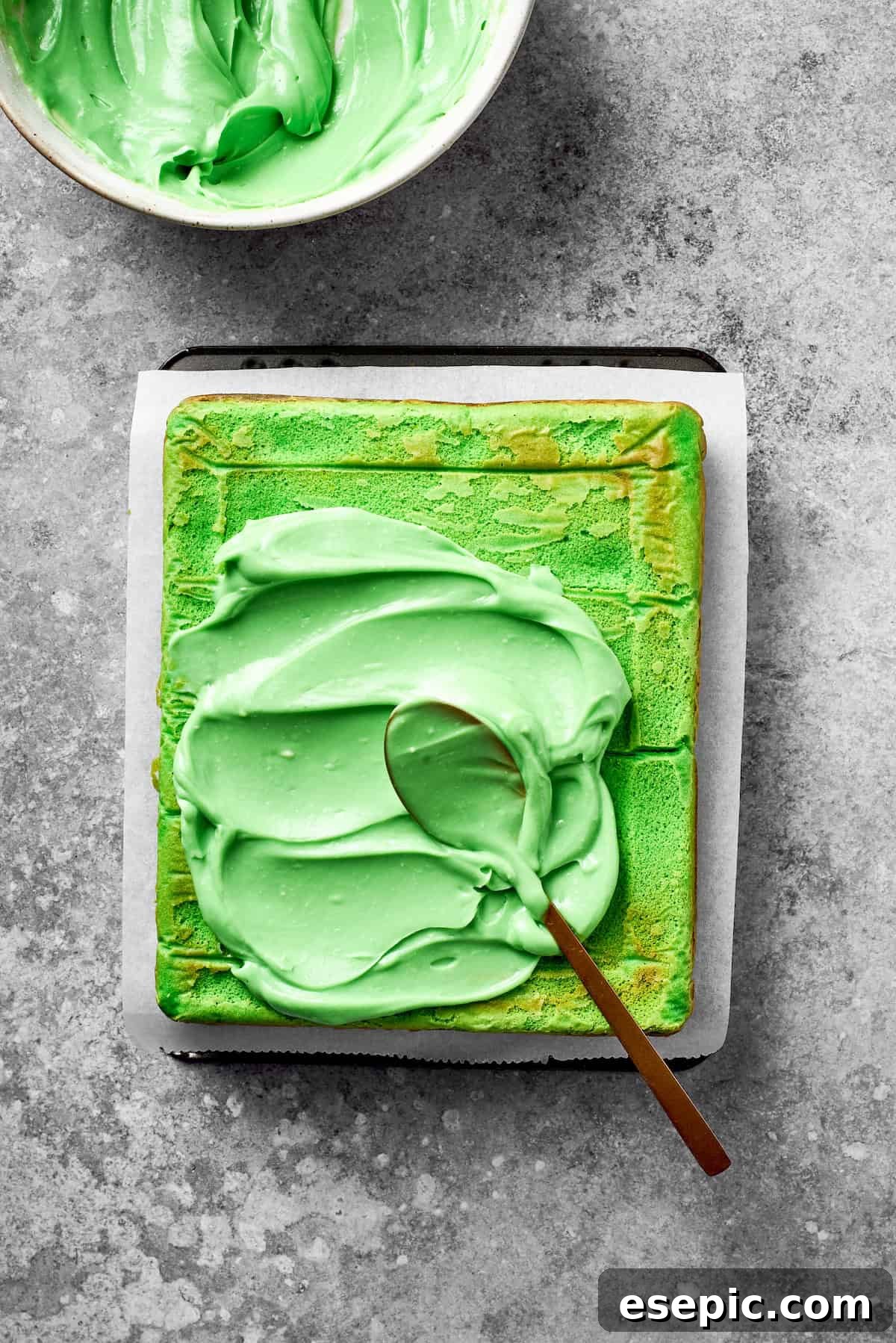 Vibrant green icing is generously spread onto the top of a baked cake, ready for decoration.