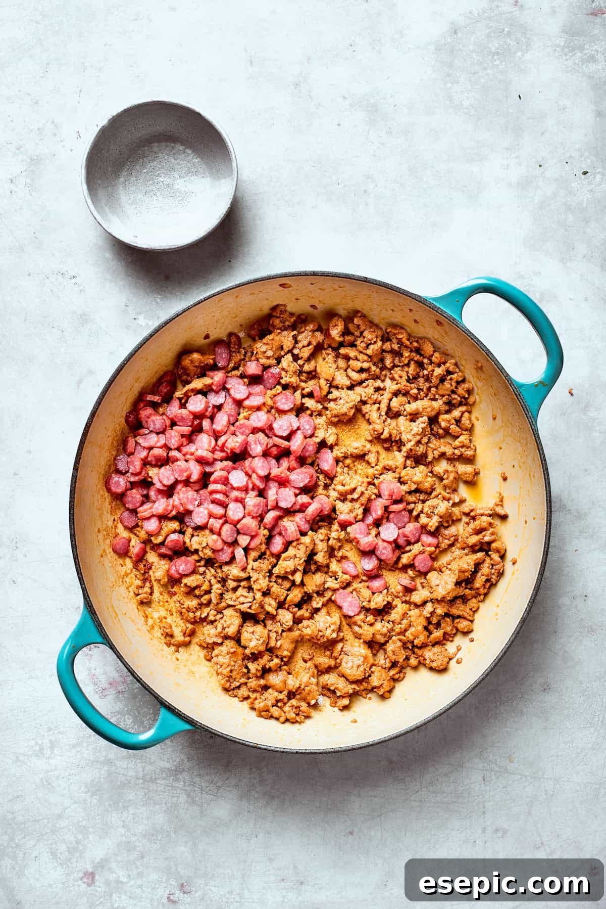 Pepperoni is added to a pan of browned sausage, showing the next step in the recipe.