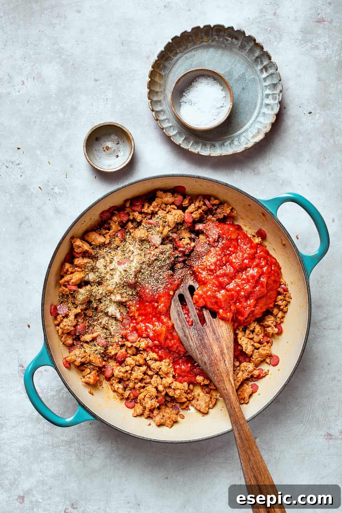 Tomato sauce is stirred into a pan with sausage and pepperoni, beginning the sauce base.