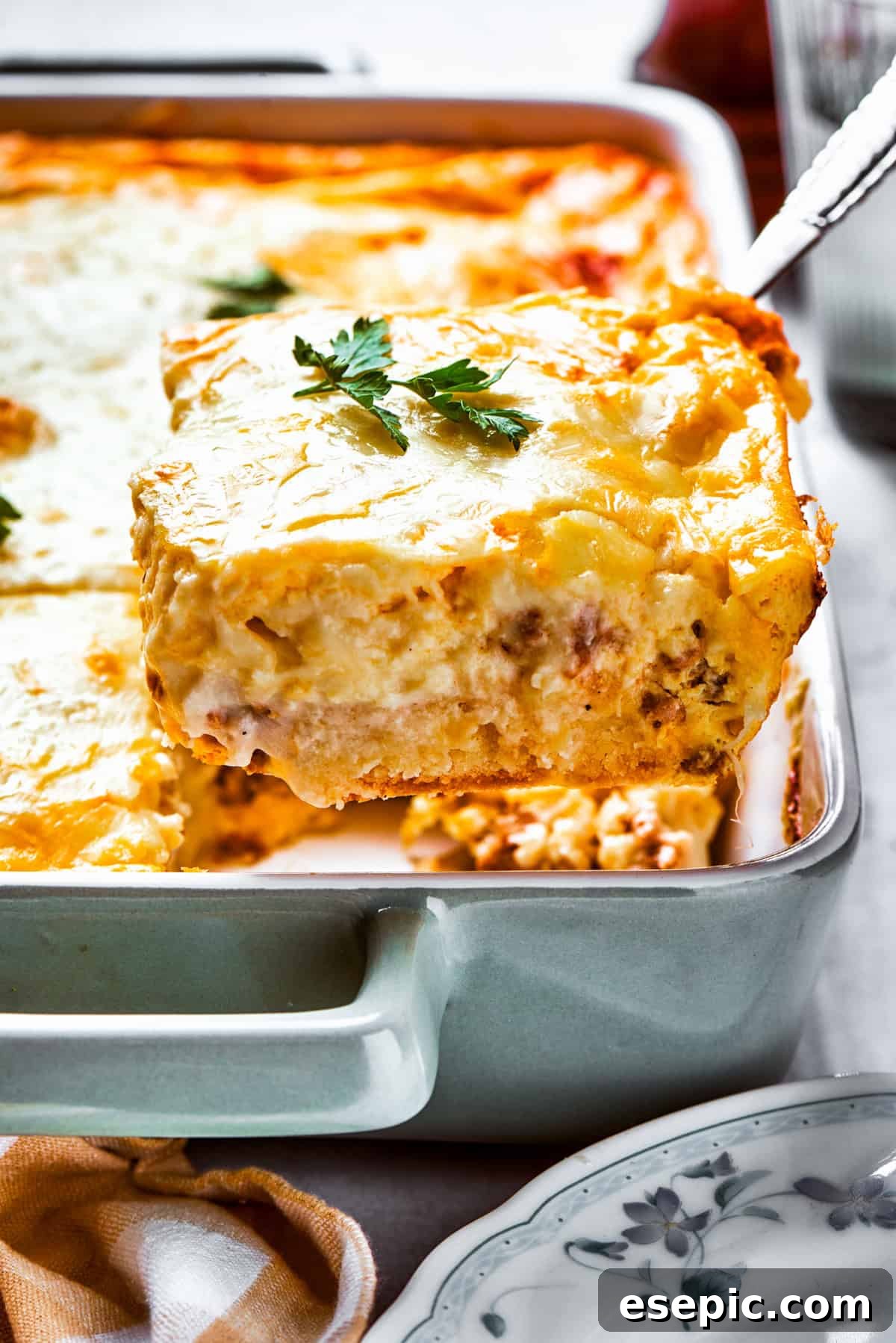 Lifting up a slice of biscuits and gravy casserole from a baking dish, showcasing layers of golden biscuits, creamy gravy, savory sausage, and melted cheese.