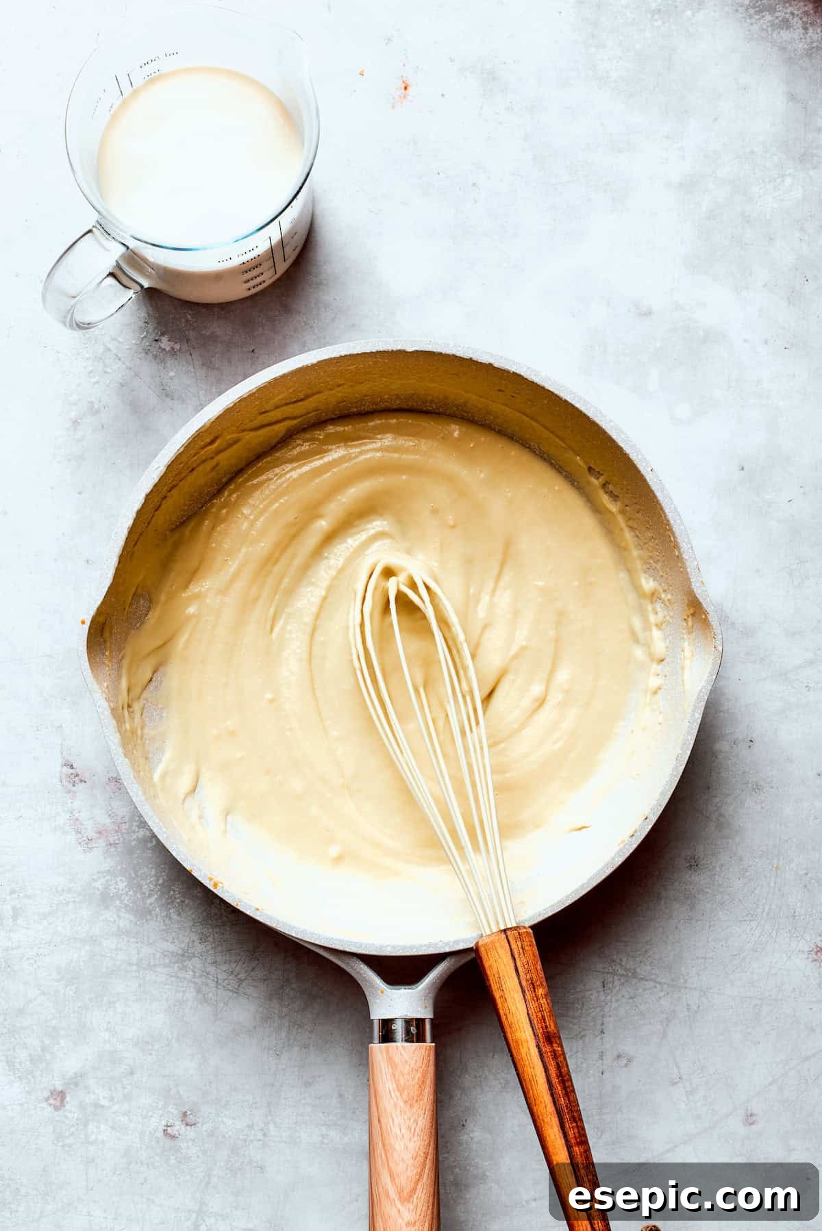 Milk being gradually whisked into the roux in a skillet, preventing lumps.
