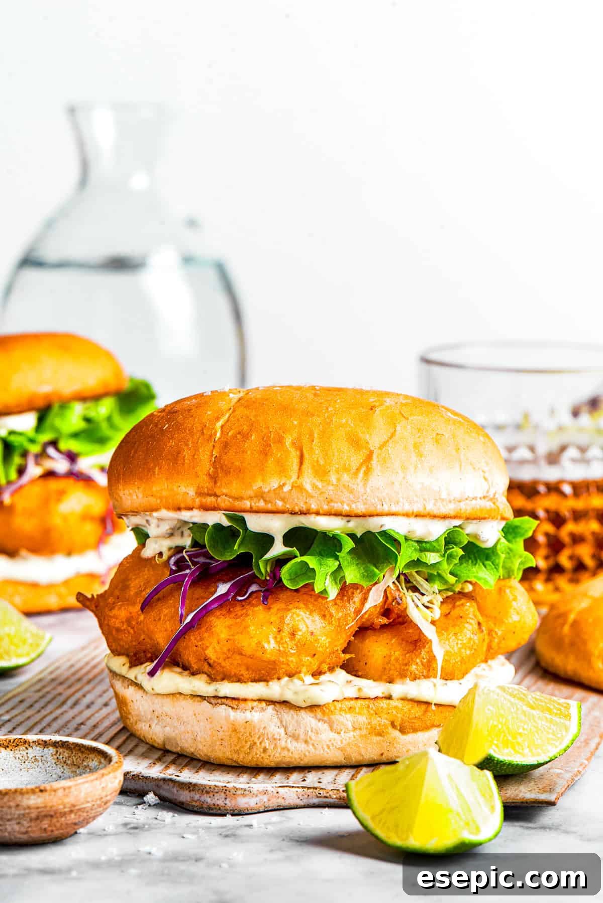 A fish sandwich on toasted buns with another sandwich and drinks placed behind it.
