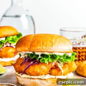 A fish sandwich on toasted buns with another sandwich and drinks placed behind it.
