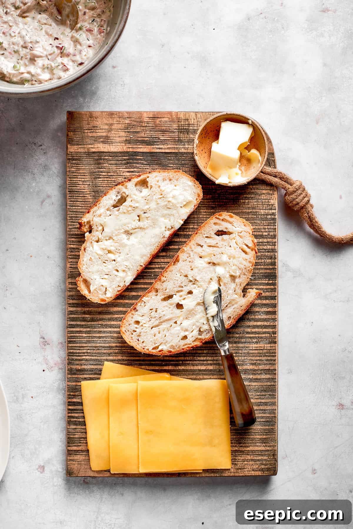 This toasty tuna melt recipe features a blend of creamy tuna salad and melty cheese stuffed between slices of sourdough and pan-fried to a crisp. Slices of sourdough bread, softened butter, and cheddar cheese slices are arranged on a cutting board, ready for sandwich assembly.