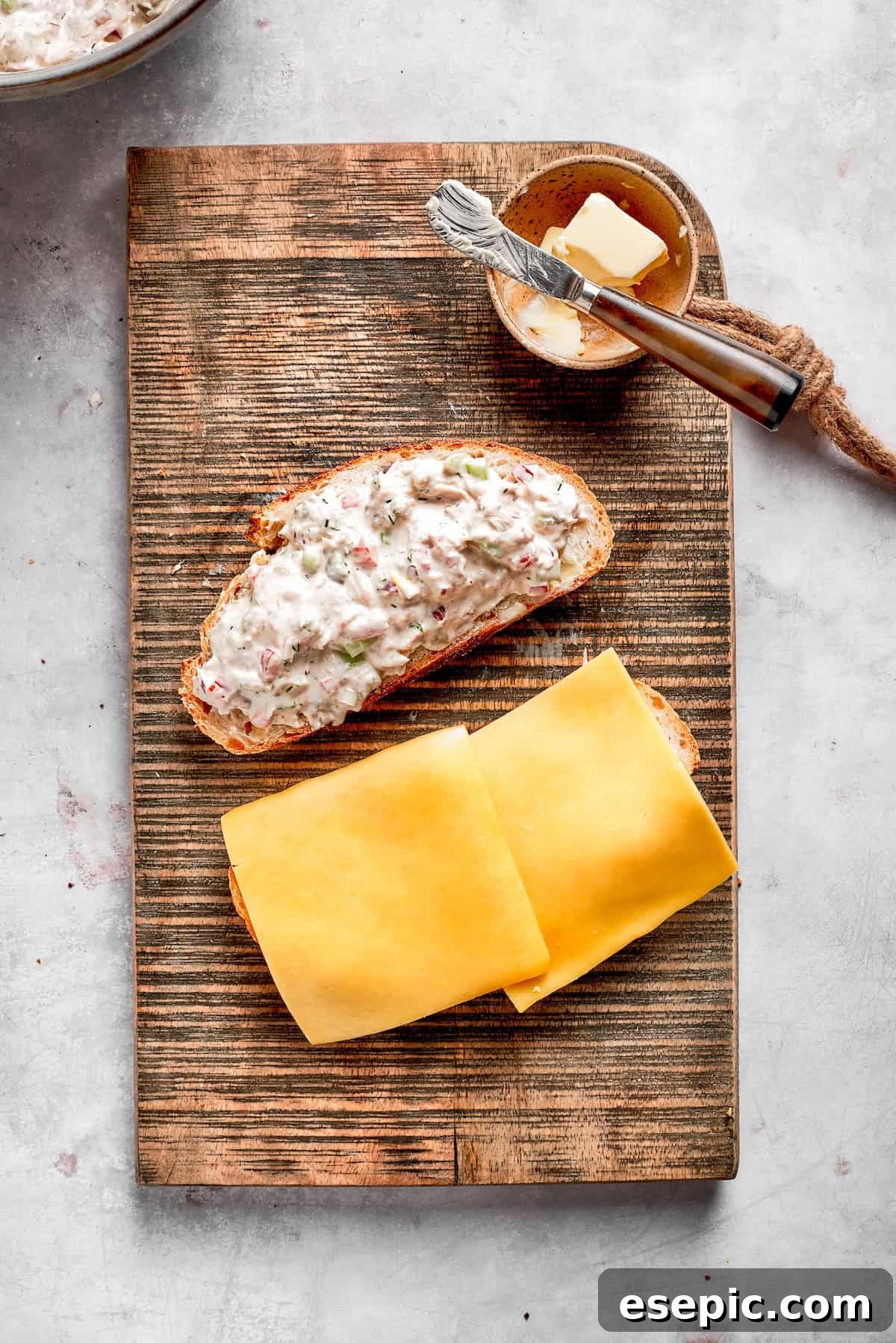 This toasty tuna melt recipe features a blend of creamy tuna salad and melty cheese stuffed between slices of sourdough and pan-fried to a crisp. Two slices of sharp cheddar cheese are placed on the unbuttered side of a slice of sourdough bread, forming the base of the tuna melt.